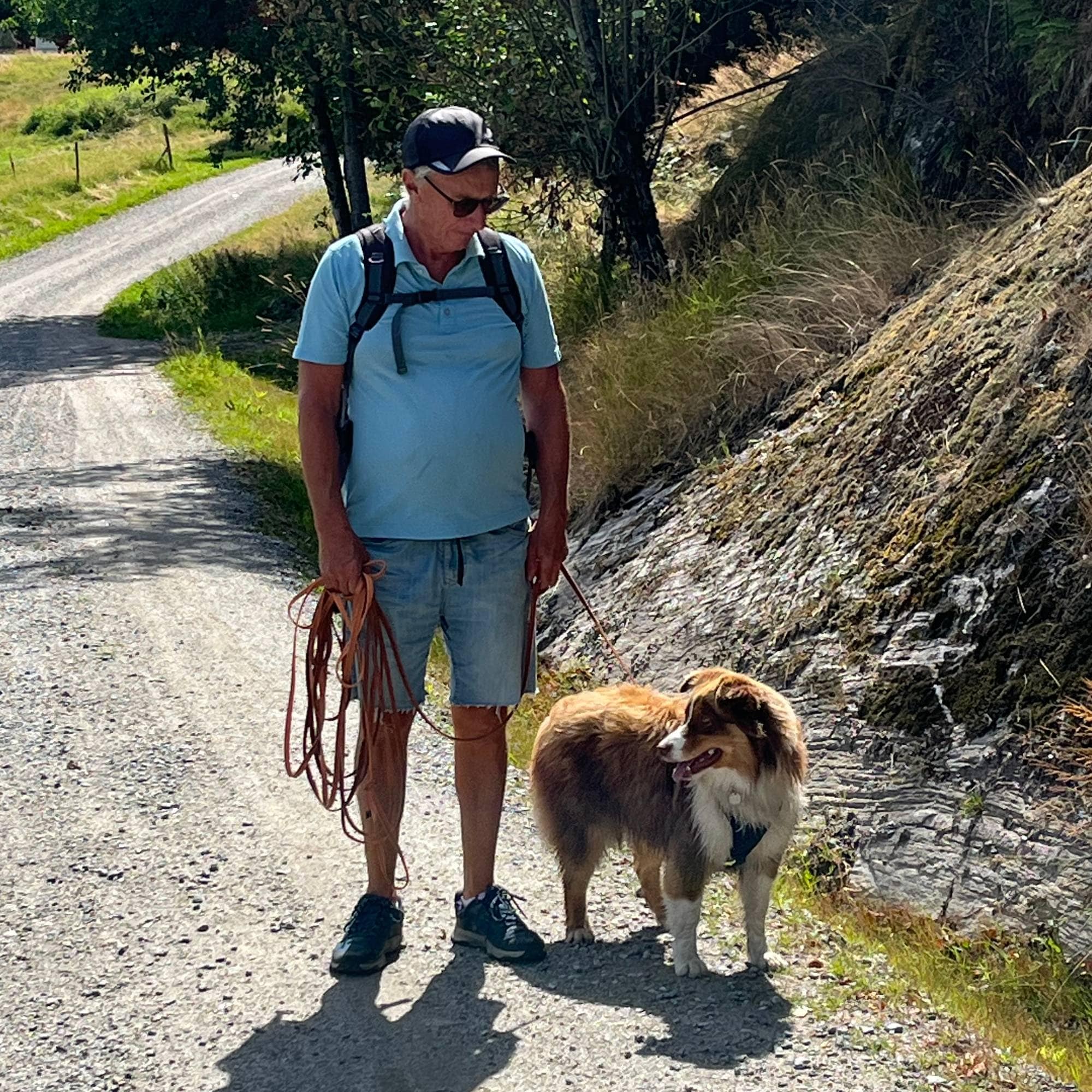 En mann med ryggsekk og hund på tur langs en grusvei i solfylt terreng. (Bildebeskrivelsen er laget av en KI-tjeneste)