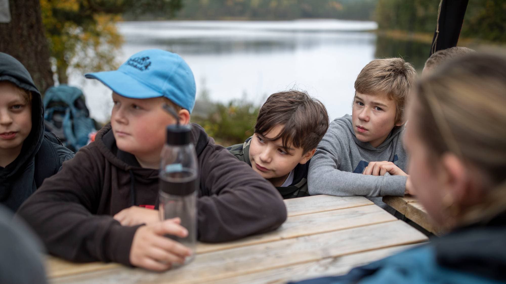 Gruppe barn sitter rundt et trebord utendørs ved et vann. (Bildebeskrivelsen er laget av en KI-tjeneste)