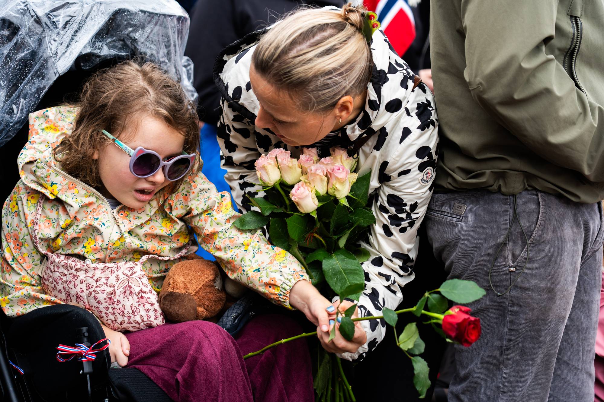 Kvinne gir en rose til jente i rullestol. Jenta holder en bamse og har solbriller. (Bildebeskrivelsen er laget av en KI-tjeneste)