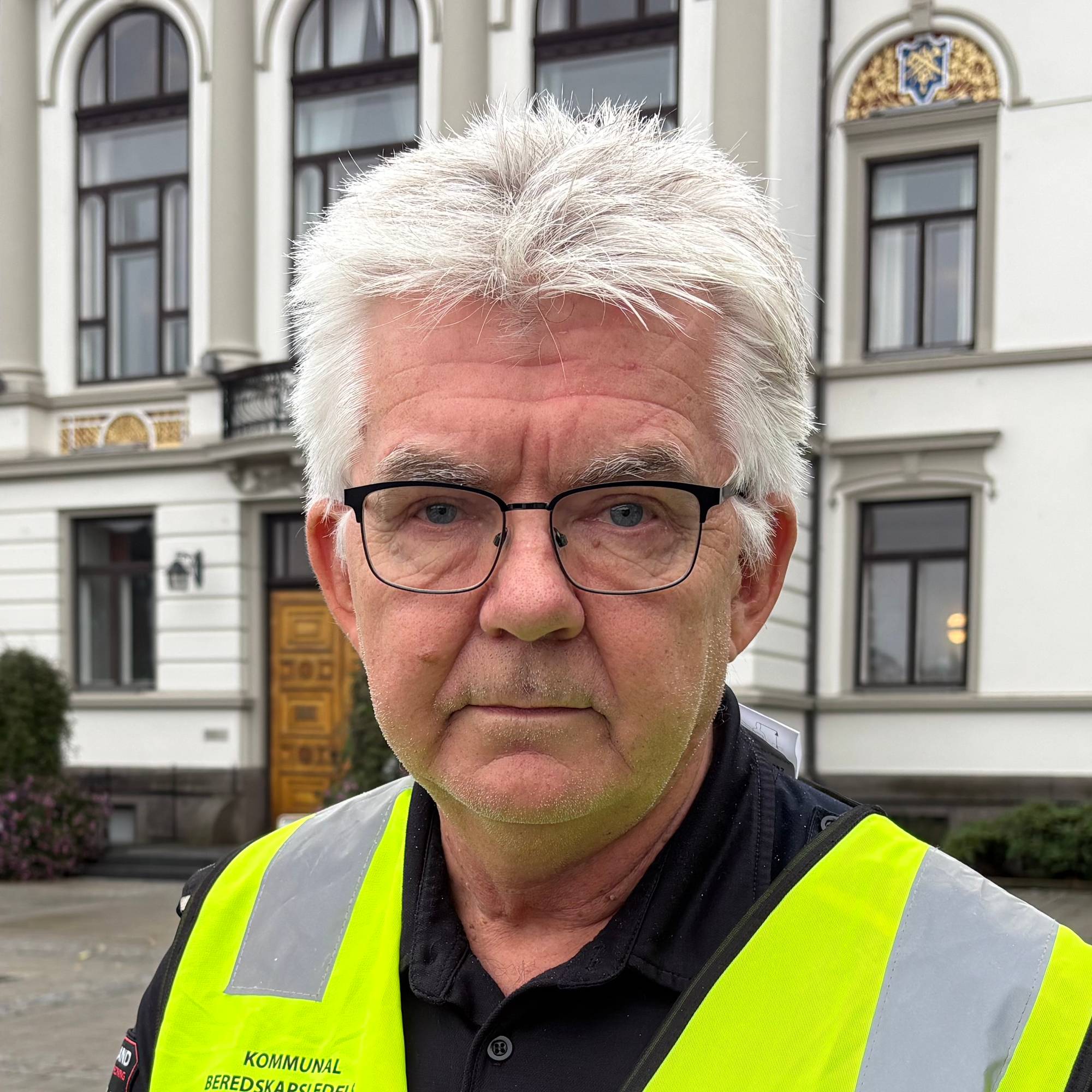 Mann i gul refleksvest med påskriften Grenlands kommunene foran en stor bygning. (Bildebeskrivelsen er laget av en KI-tjeneste)
