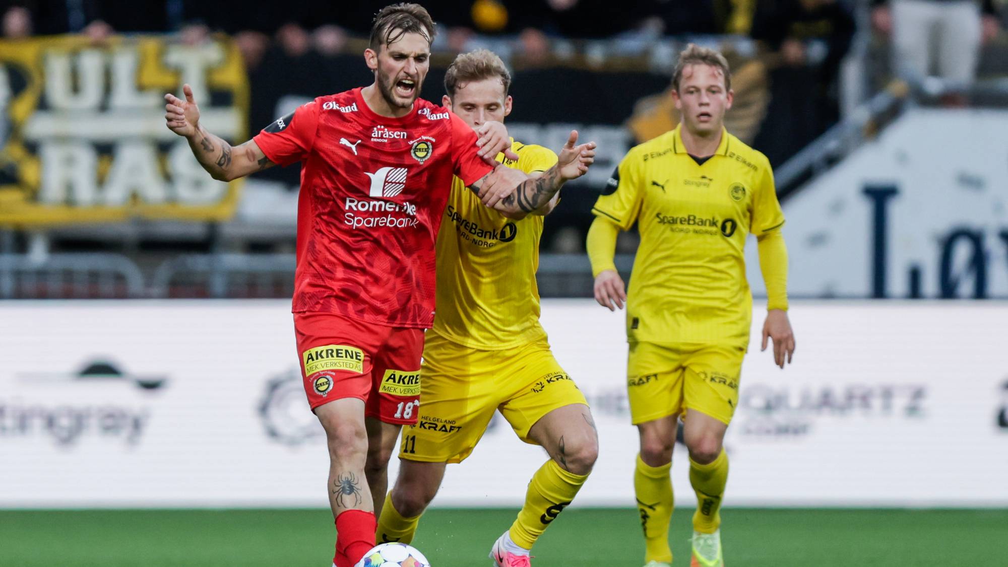 Fotballspiller i rød drakt kjemper om ballen mot en motspiller i gult på en gressbane. (Bildebeskrivelsen er laget av en KI-tjeneste)