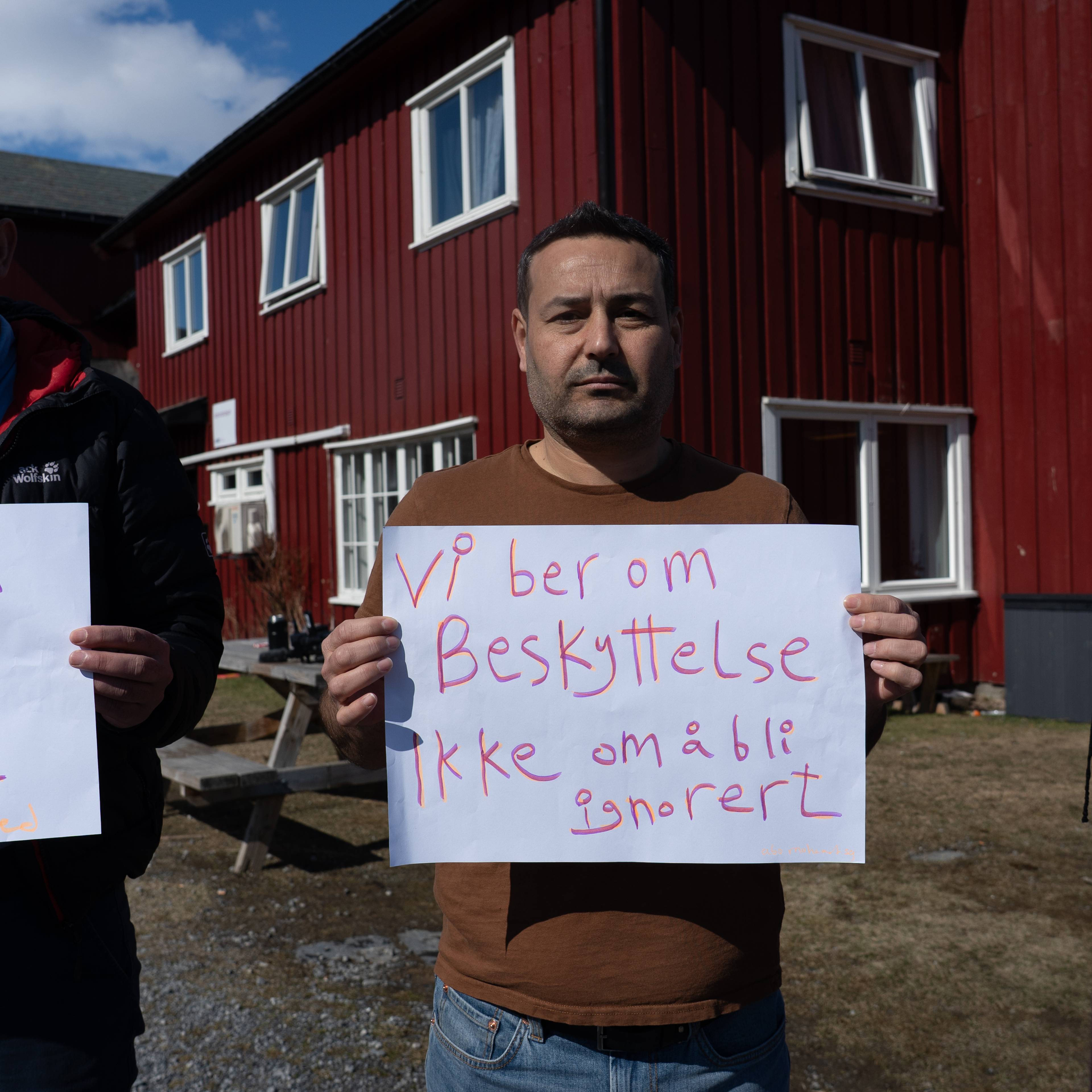 Menn holder skilt framfor ei raud bygning. Et skilt har teksten "Våre barn fortener ei betre framtid". Et anna skilt har teksten "Vi ber om Beskyttelse Ikke om å bli ignorert". Ei kvinne med hijab står til høyre. (Bildebeskrivelsen er laget av ei KI-teneste)