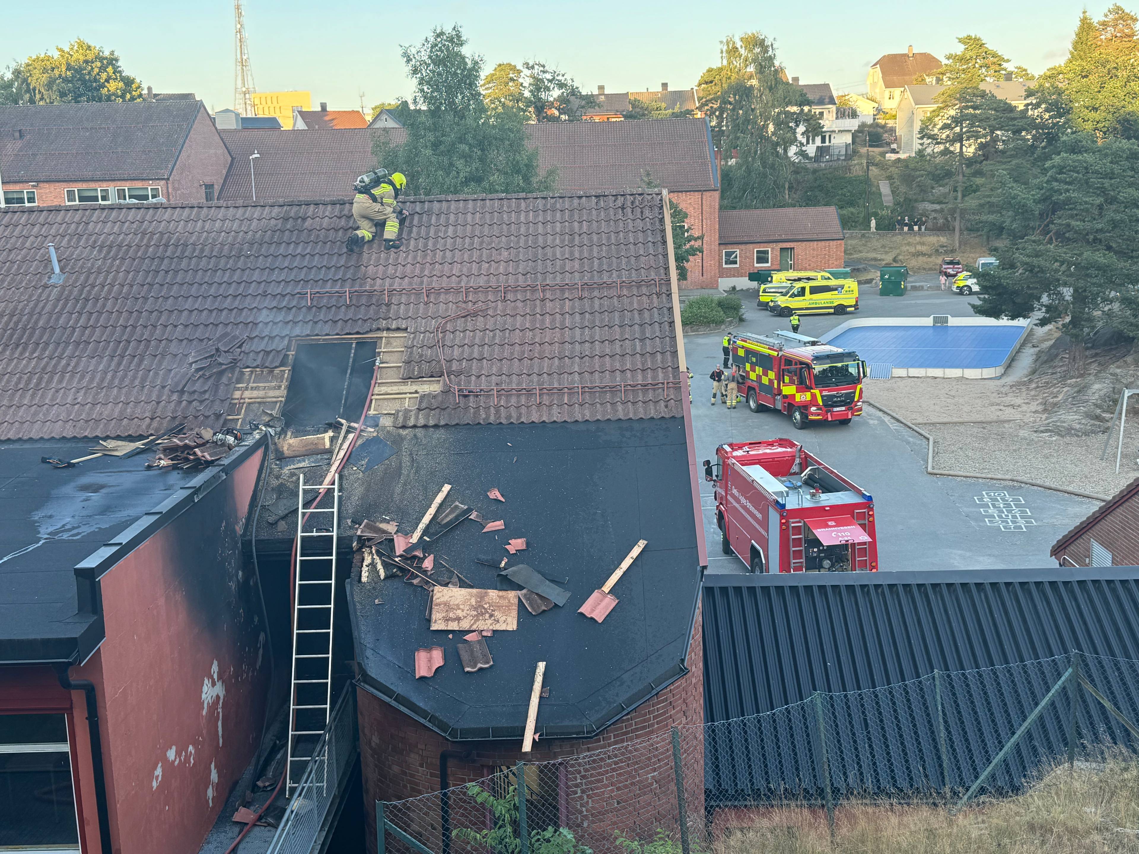 Brannvesenet arbeider på taket av en bygning etter brann. Nødetater er til stede på plassen. (Bildebeskrivelsen er laget av en KI-tjeneste)