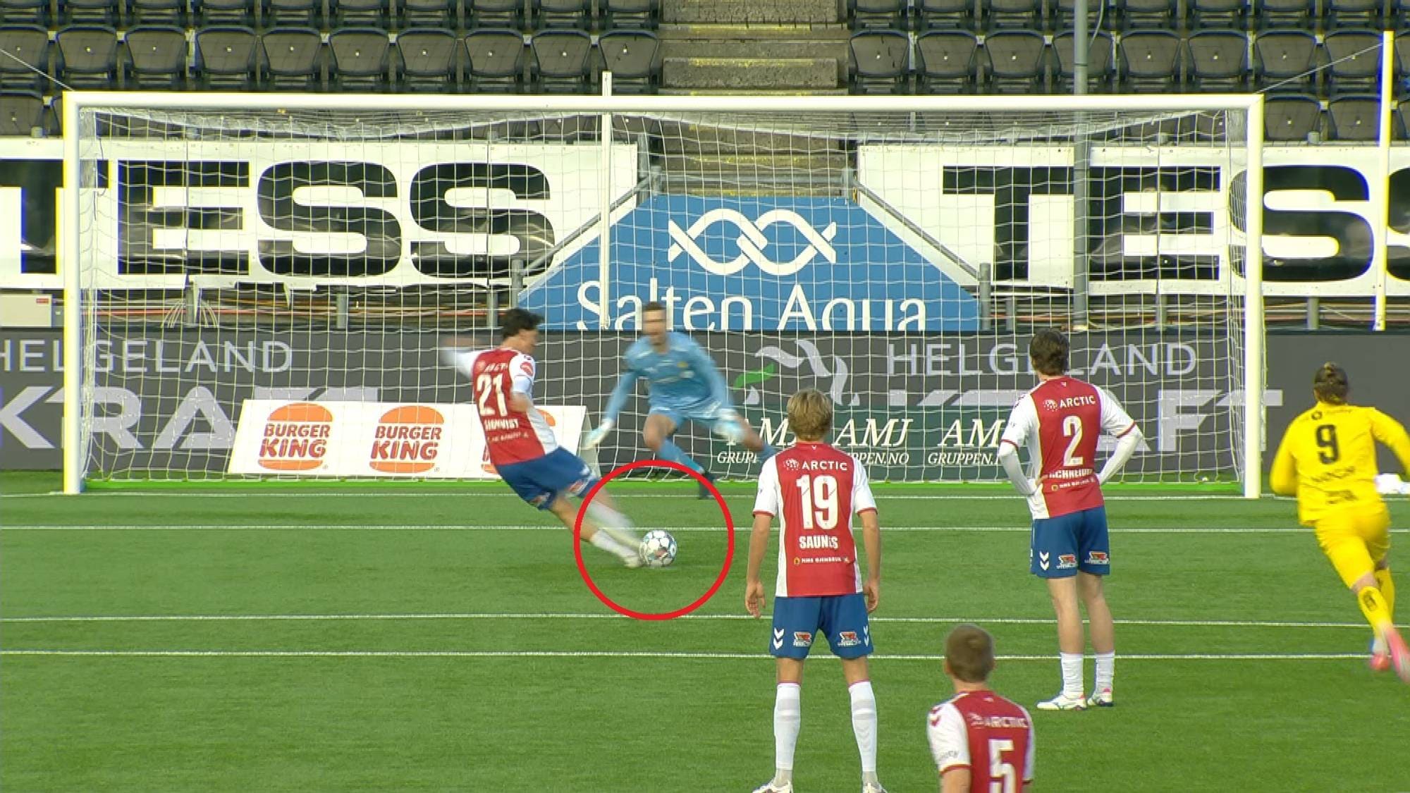 En fotballspiller i rødt og blått er i ferd med å sparke ballen mot mål. En keeper i lyseblå drakt er klar til å redde. Andre spillere i rødt og en i gult er også på banen. Bak målet er det reklameplakater. (Bildebeskrivelsen er laget av en KI-tjeneste)