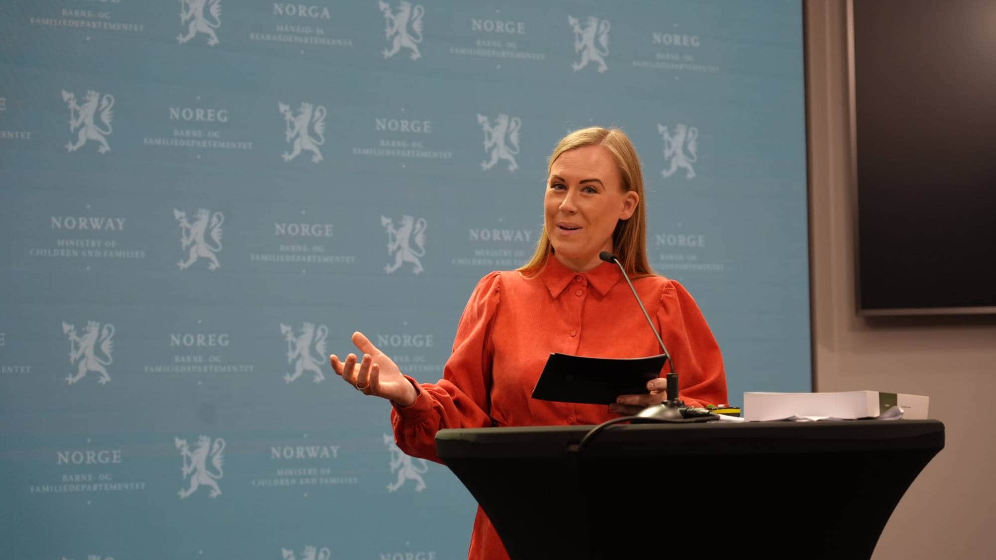 En kvinne i oransje kjole holder en tavle og snakker ved et podium. Bak henne er et blått bakteppe med gjentatte logoer fra Barne- og familiedepartementet. Hun gestikulerer med høyre hånd. (Bildebeskrivelsen er laget av en KI-tjeneste)