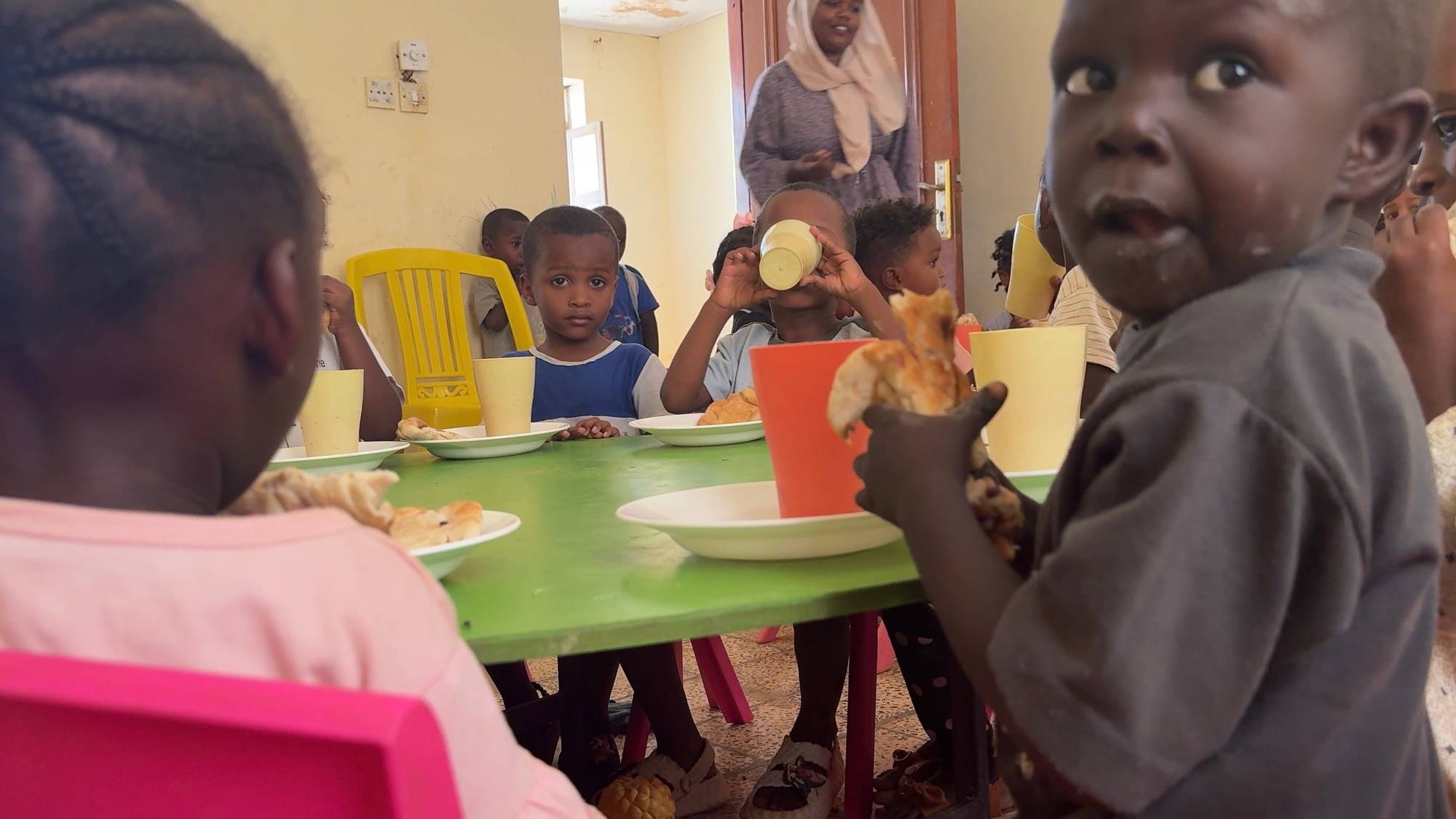 Afrikanske barn sit rundt eit bord. Dei et brød, drikker frå ein kopp og kikkar nysgjerrig mot kamera. 