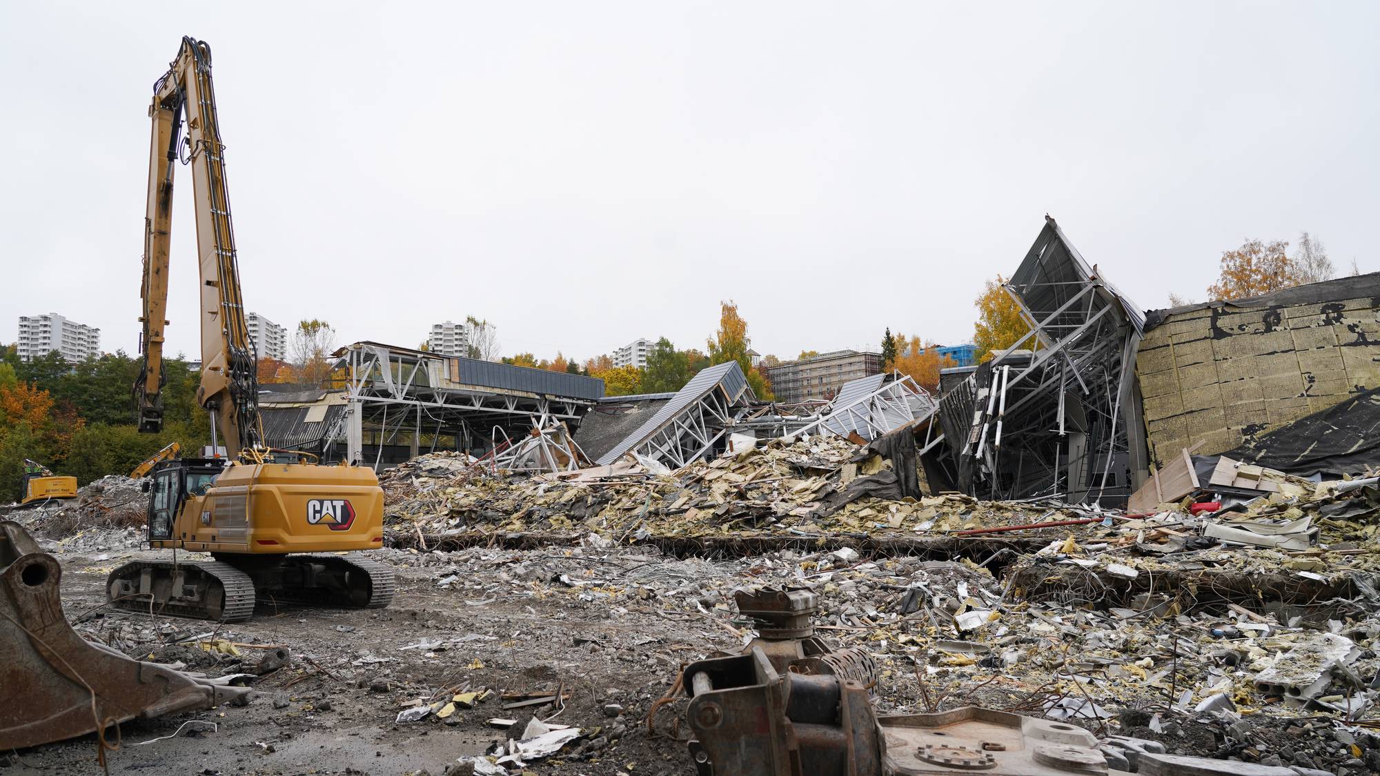 En gravemaskin står foran en bygning under riving. Ruiner og bygningsrester dekker tomten. (Bildebeskrivelsen er laget av en KI-tjeneste)
