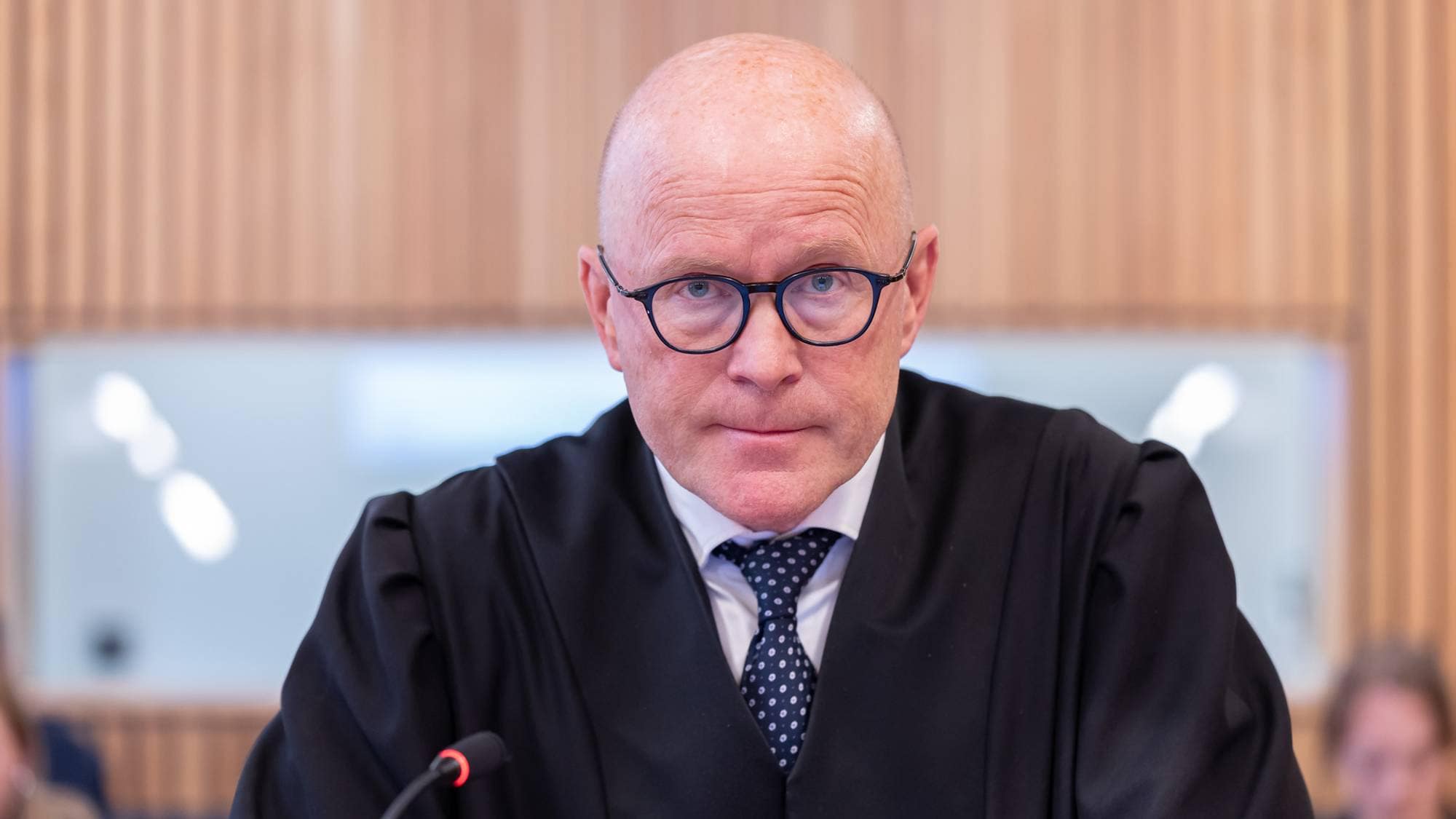 En mann iført svart kappe og blått slips står ved en benk i en rettssal. mannen har brille med mørk innfatning. Han har ikke hår på hodet. Mannen som er aktor kikker strengt og konsentrert på fotografen