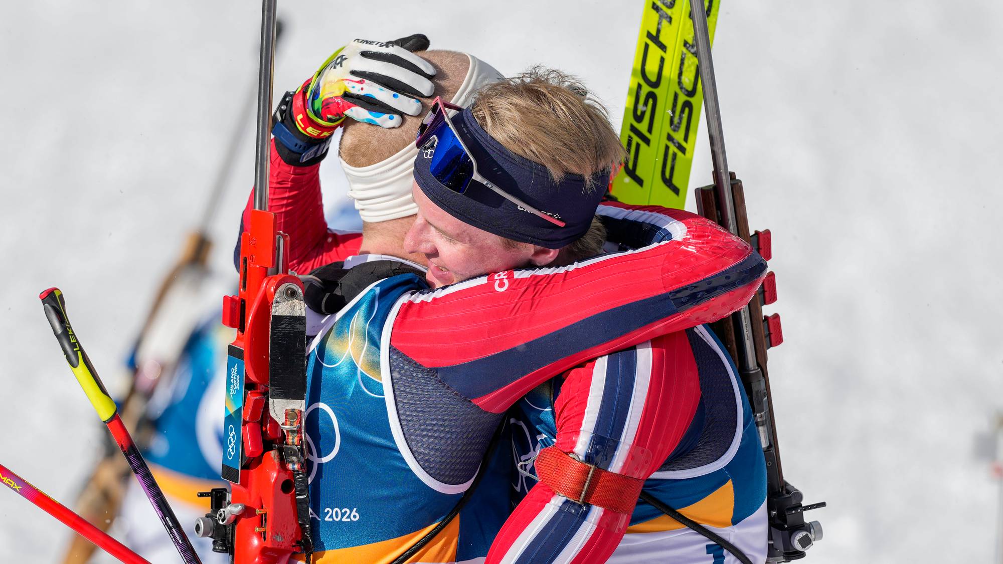 To skiskyttere i norske landslagsdrakter omfavner hverandre i et snødekket landskap. (Bildebeskrivelsen er laget av en KI-tjeneste)