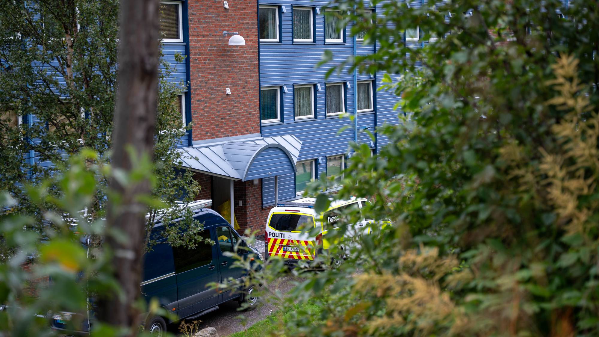 Politibil og varebil parkert utenfor en boligblokk. Grønne trær rammer inn motivet. (Bildebeskrivelsen er laget av en KI-tjeneste)