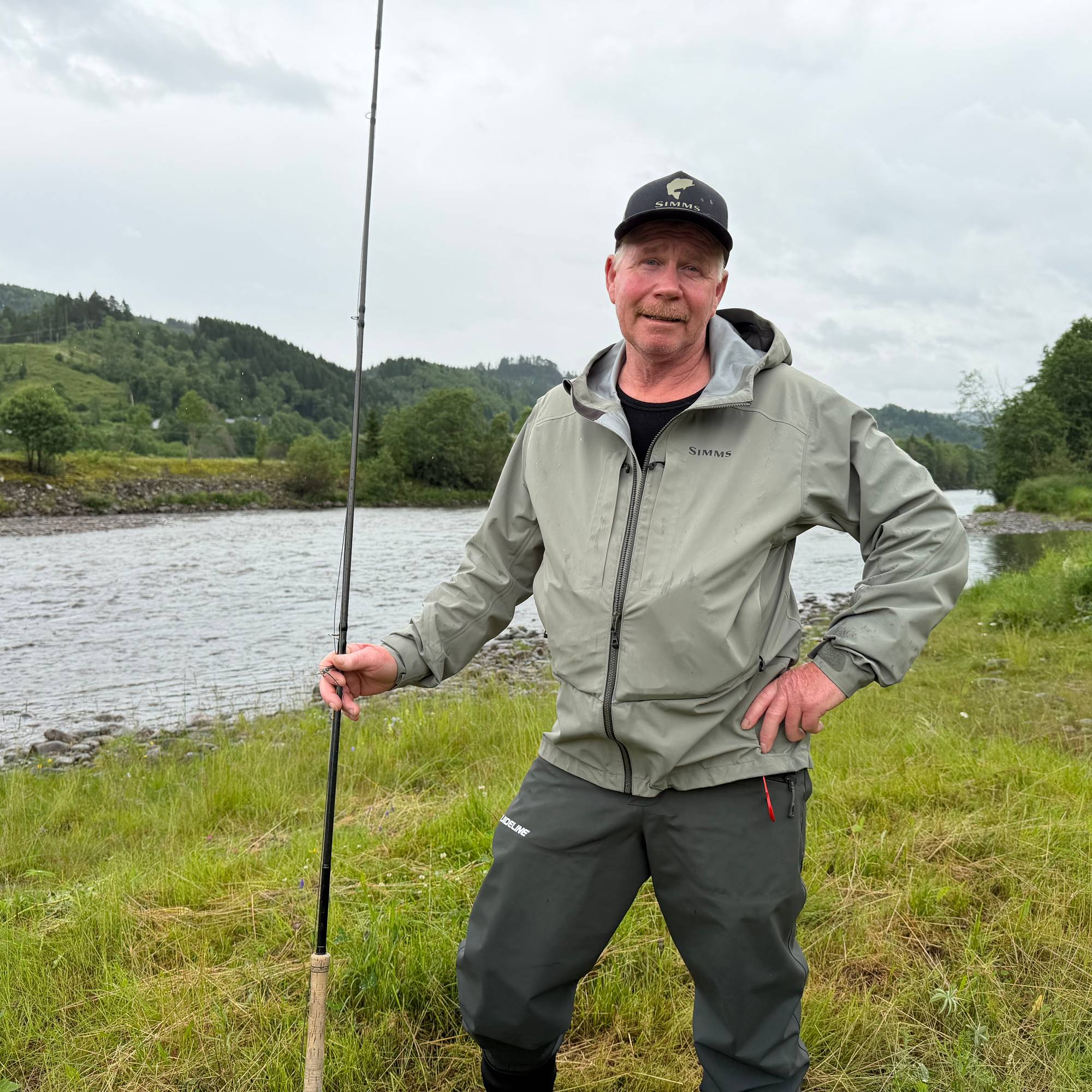 Mann med fiskestang står ved en elvebredd. Bakgrunn med skog og fjell. (Bildebeskrivelsen er laget av en KI-tjeneste)