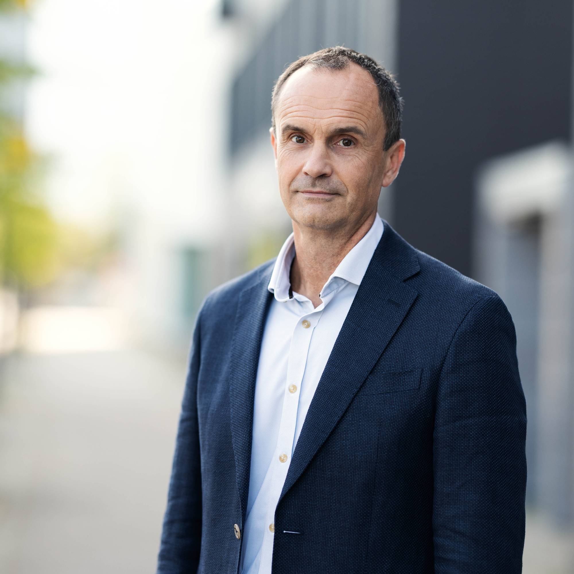En mann i lys blå skjorte og blå dressjakke ser alvorlig inn i kamera. Bakgrunnen er skurrete