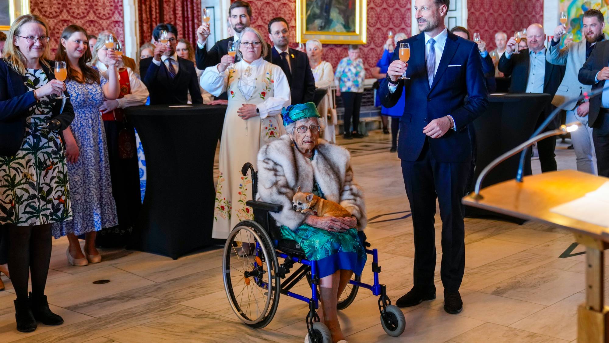 Prinsesse Astrid - En forsamling med pyntede mennesker, prinsesse Astrid i rullestol foran, med turkis hatt og kjole og hvitt sjal