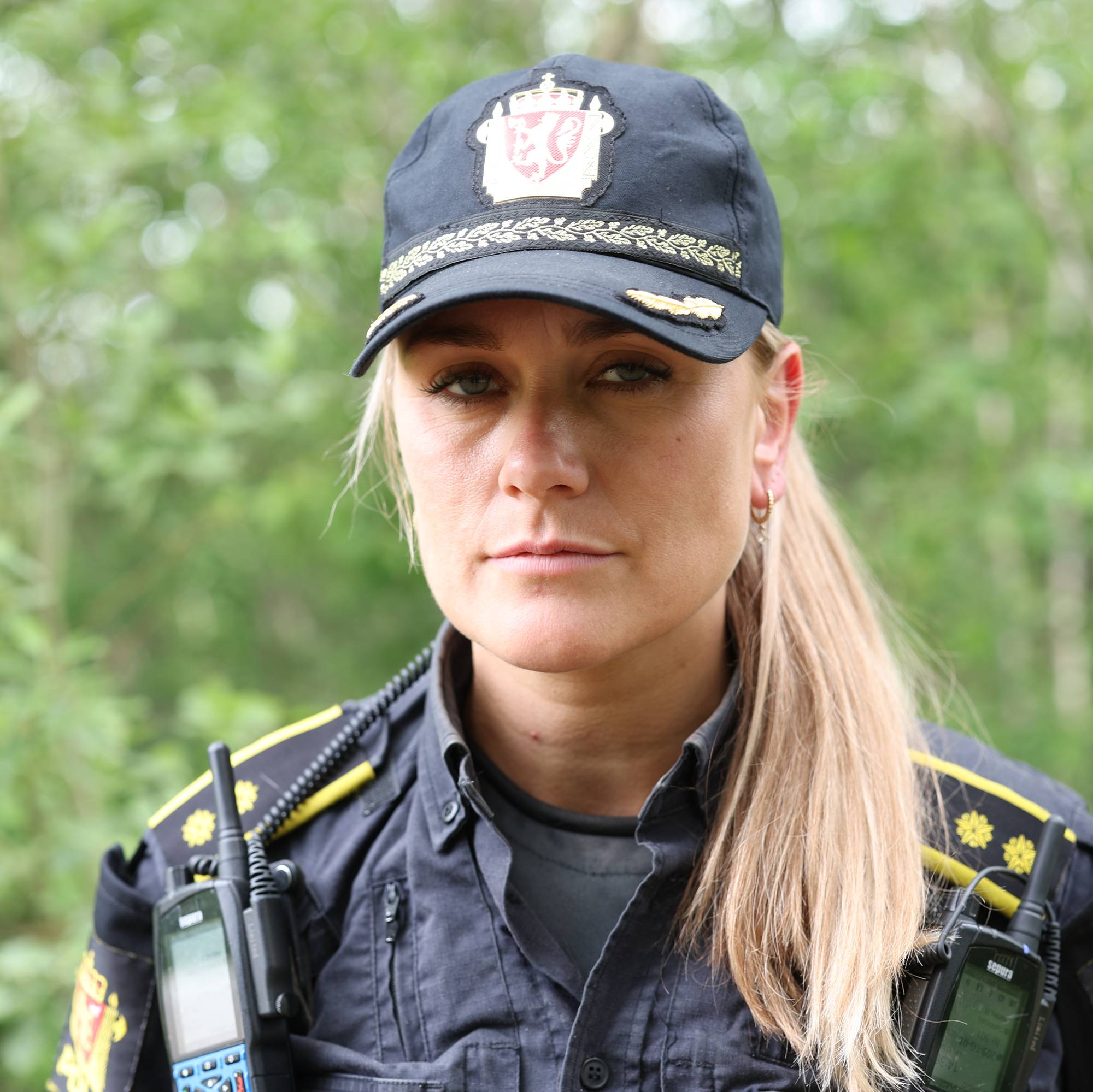 En kvinne i politiuniform med caps ser mot kameraet. Hun har to radiokommunikasjonsenheter festet til uniformen. Bak henne er det grønne trær og vegetasjon. Bildet er et nærbilde av kvinnen. (Bildebeskrivelsen er laget av en KI-tjeneste)