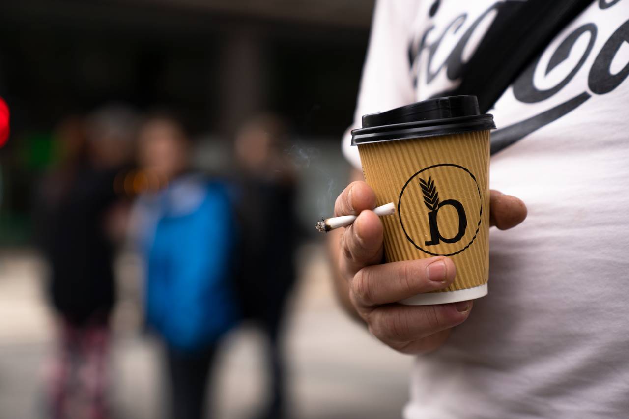 En rusbruker holder et brunt kaffekrus i hånden samt en sigarett. i bakgrunnen ser vi noen andre rusbrukere.