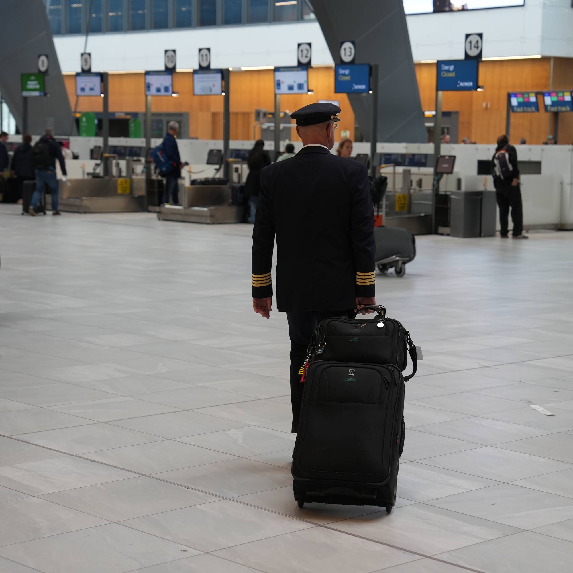 En pilot går med trillekoffert gjennom en flyplasshall med innsjekkingsautomater i bakgrunnen. 