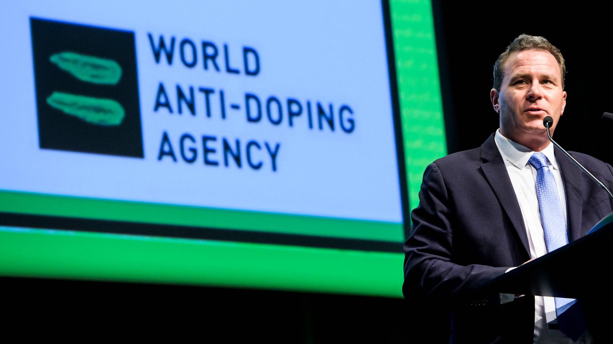 En mann i dress snakker ved et podium. Bak ham vises logoen til World Anti-Doping Agency. Bakgrunnen er mørk med en grønn stripe langs bunnen. Mannen holder et dokument i hånden. (Bildebeskrivelsen er laget av en KI-tjeneste)