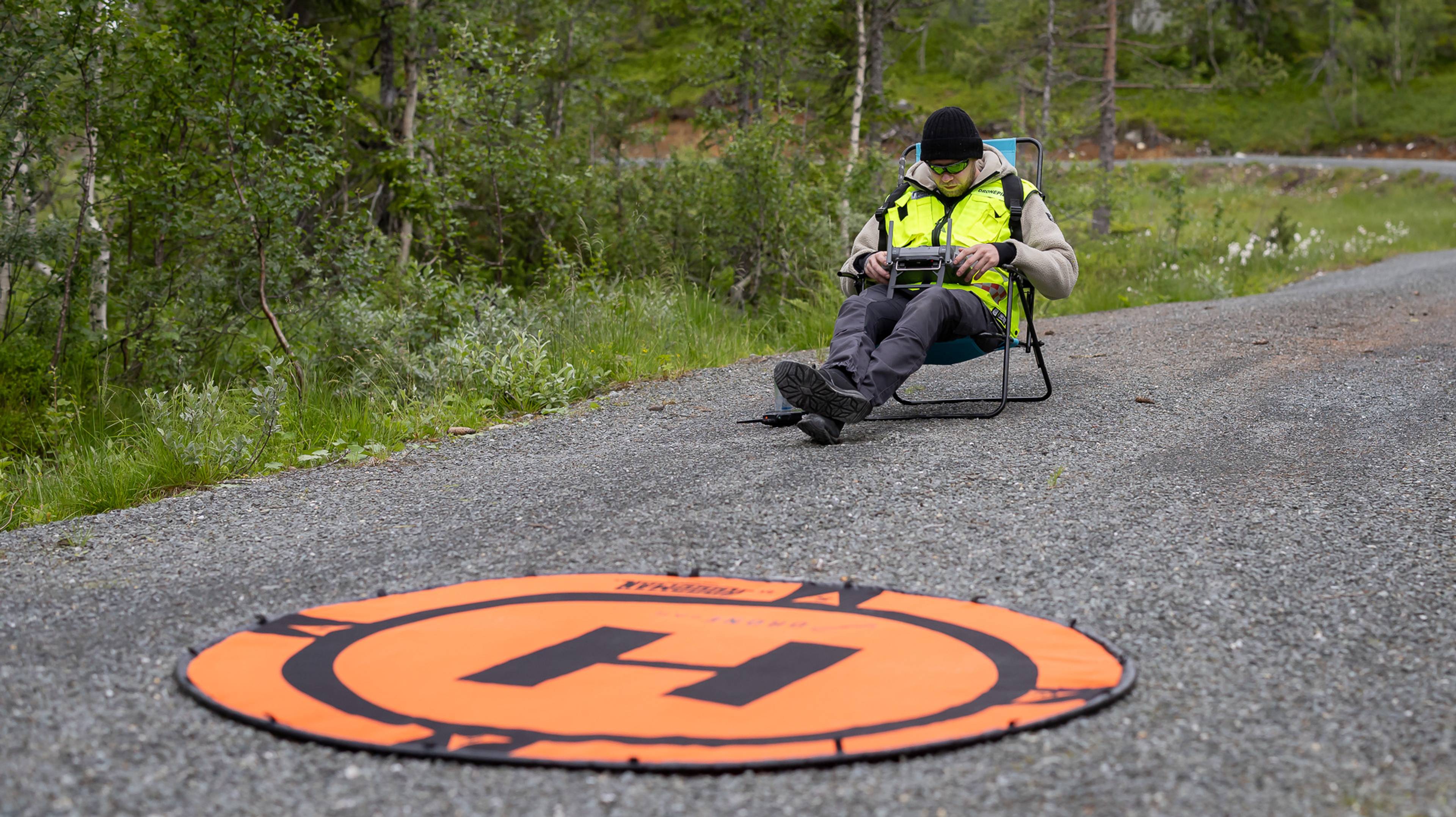 En person sitter i en fluktstol og styrer en drone. Han sitter på en grusvei. Foran ham ser vi en stor matte liggende på bakken. En stor H på matten er godt synlig
