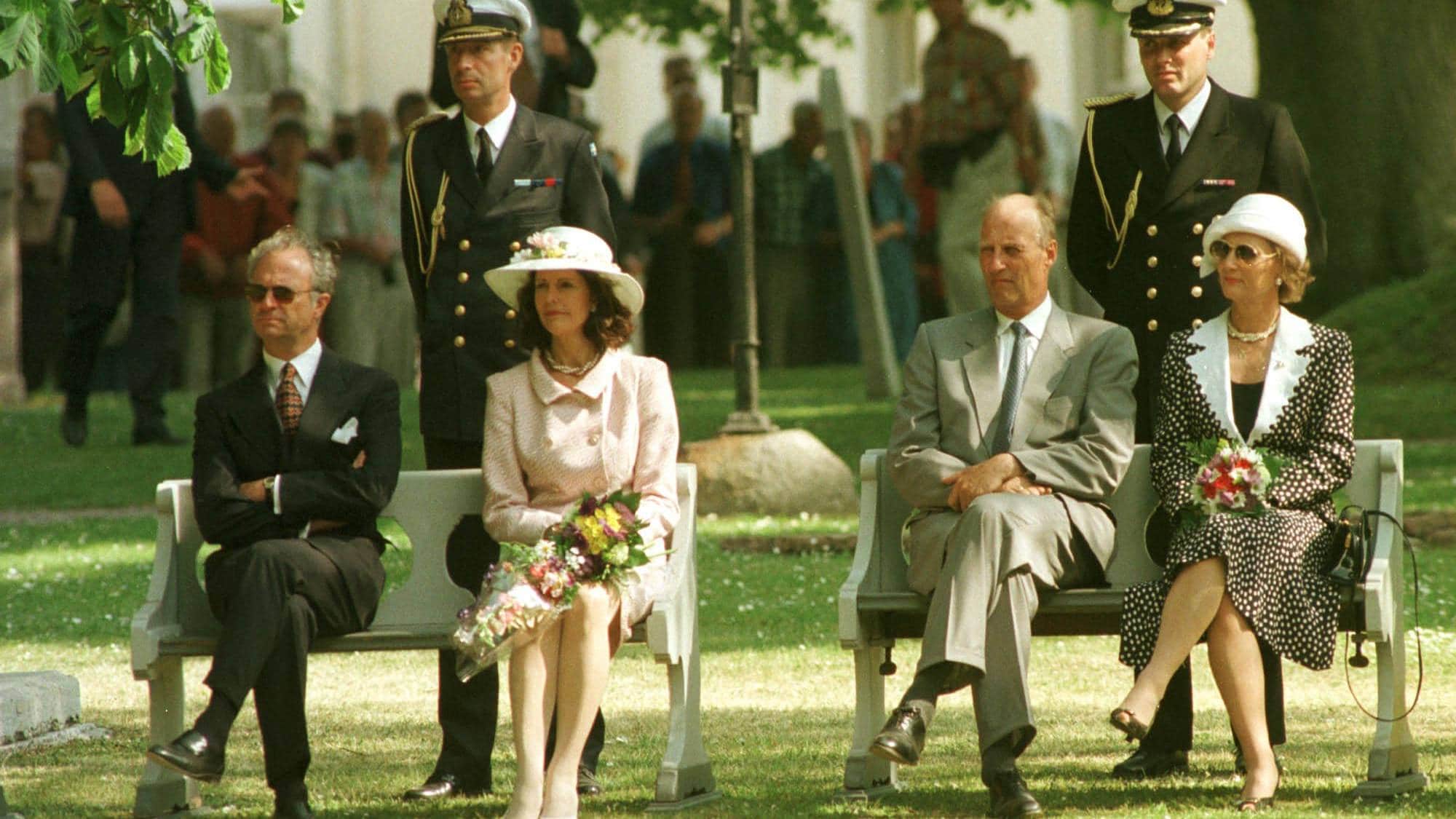 Kong Harald og dronning Sonja sitter på en benk i det fri. De er omgitt av folk i uniform og sivile antrekk. To menn i mørke uniformer står bak kongen og dronningen. (Bildebeskrivelsen er laget av en KI-tjeneste)