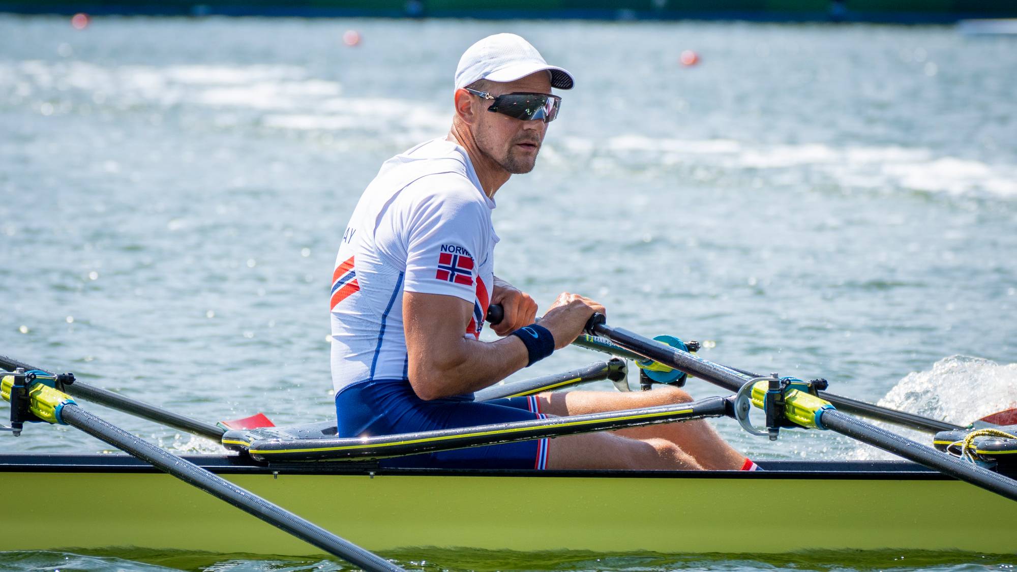 En mann i en robåt ror på vannet. Han har på seg en hvit t-skjorte med det norske flagget på ermet og blått tøy. Bakgrunnen viser vann og en grønn plakat med "TOKYO 2020". Han har på seg solbriller og en caps. (Bildebeskrivelsen er laget av en KI-tjeneste)