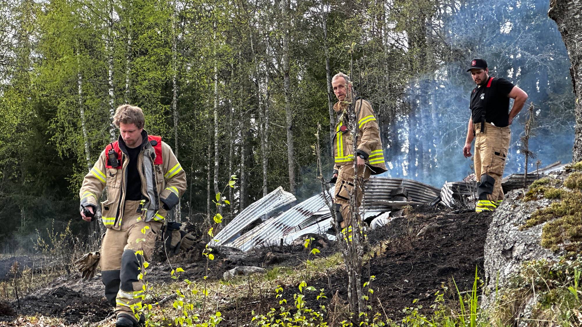 Tre brannmenn står i et nedbrent område med vrakrester av metall. De er kledd i brannverndresser. Bak dem ses et skogholt. Det ligger røyk i luften. (Bildebeskrivelsen er laget av en KI-tjeneste)