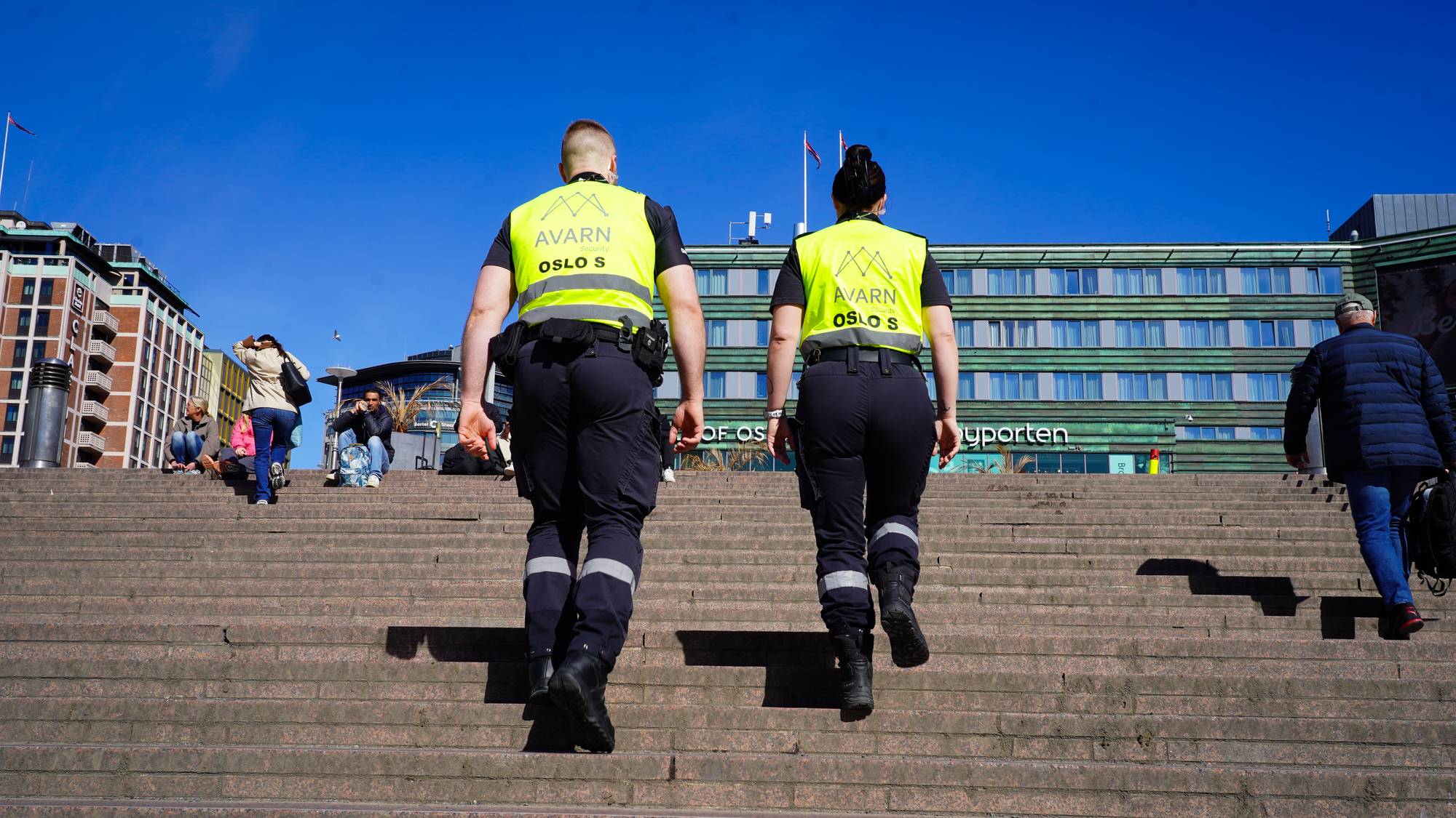 To vektere går opp en steintrapp med en bygning i bakgrunnen. Vekterne har på seg gule synlighetsskjorter med teksten "AVARN OSLO S" og mørke bukser. Flere personer sitter på trappen, og en mann går opp trappen til høyre. (Bildebeskrivelsen er laget av en KI-tjeneste)