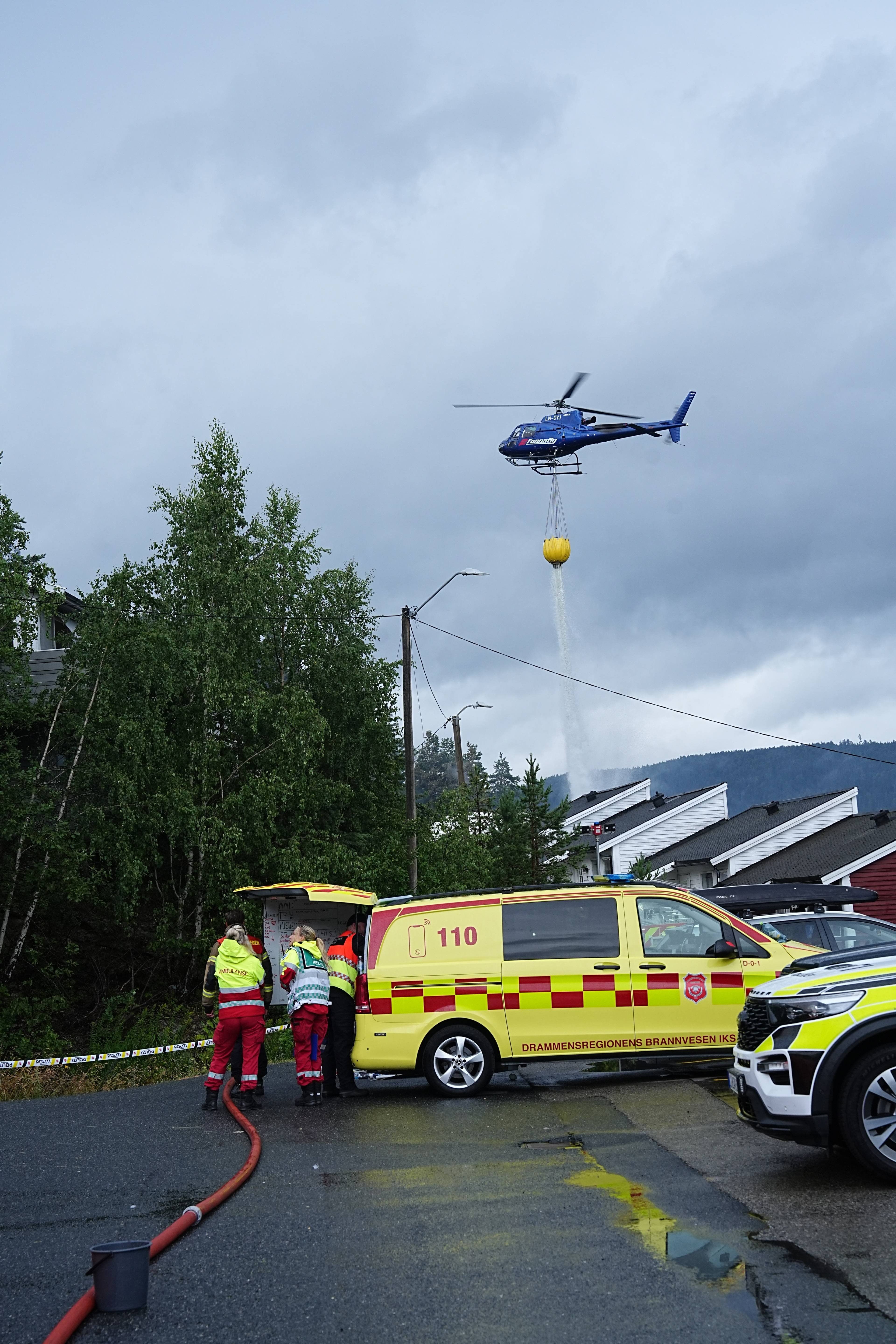 Brannhelikopter slipper vann over boligområde mens nødetater står ved en brannbil. (Bildebeskrivelsen er laget av en KI-tjeneste)