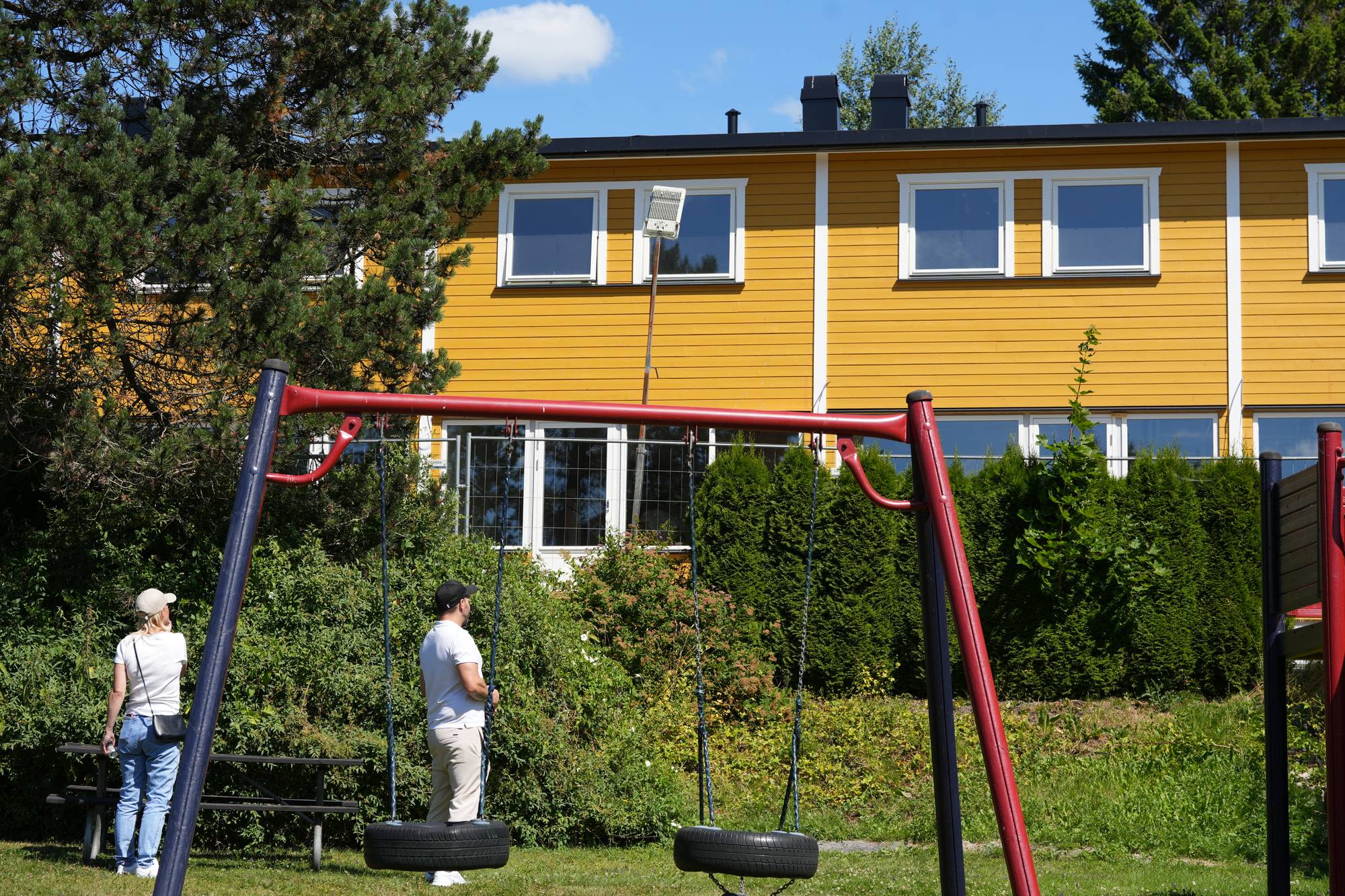 To personer står ved en lekeplass med husker foran en gul boligblokk. (Bildebeskrivelsen er laget av en KI-tjeneste)