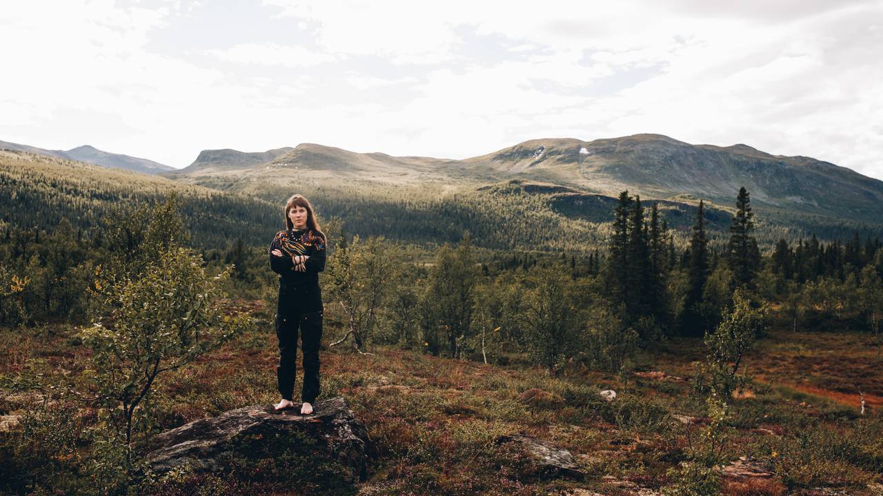 Katarina Barruk står med armene foldet på en stein og ser inn i kamera. Bak henne ser man all den herligheten som er naturen i hjemområdet hennes.
