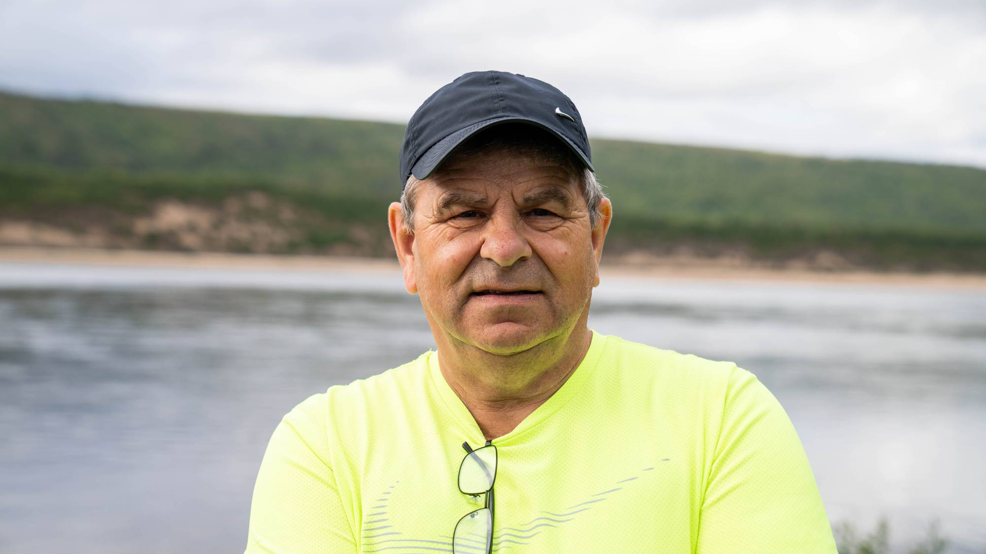 Benn Larsen, leder i Tanavassdragets fiskeriforvaltning (TF) står å ser mot kameraet forran Tanavassdraget, i en svart caps og signalgul t-skjorte.