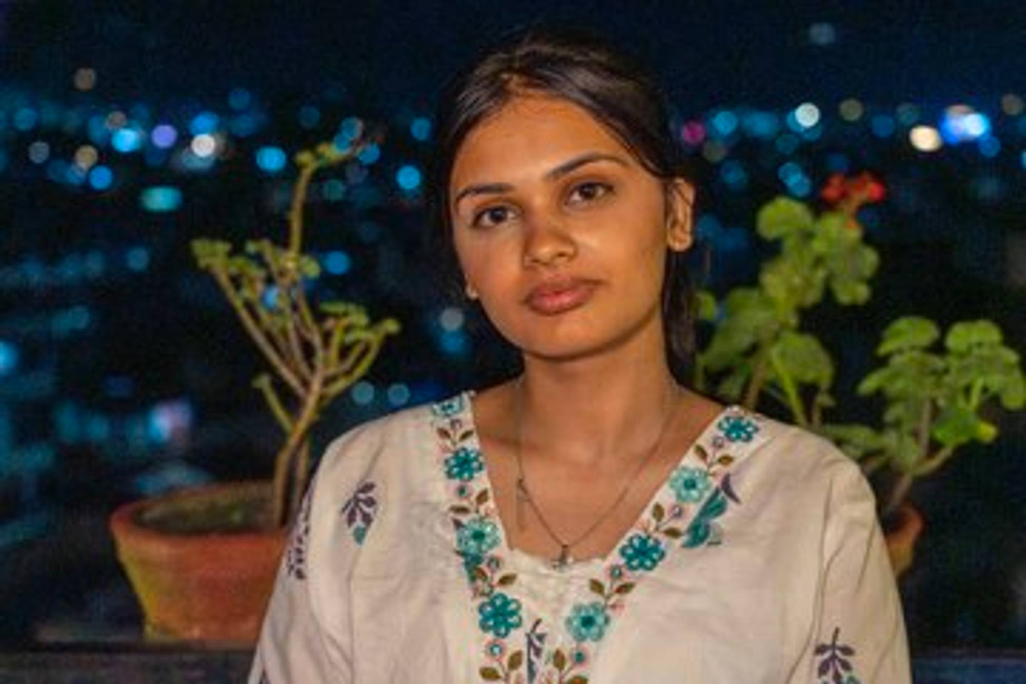 Kritika Bajgain, Katmandu - Kvinne med store brune øyne og mørkt hår strøket bak ørene, hvit skjorte med turkise blomster. Kveld med lysene fra byen i bakgrunnen. 