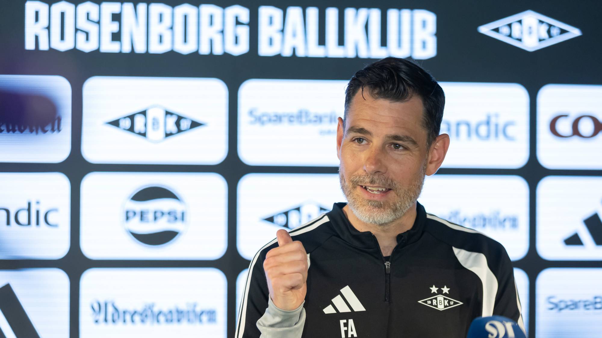 RBKs nye trener Freyr Alexandersson holder pressekonferanse. Han løfter hånden. I bakgrunnen kan vi se logo og teksten Rosenborg Ballklub
