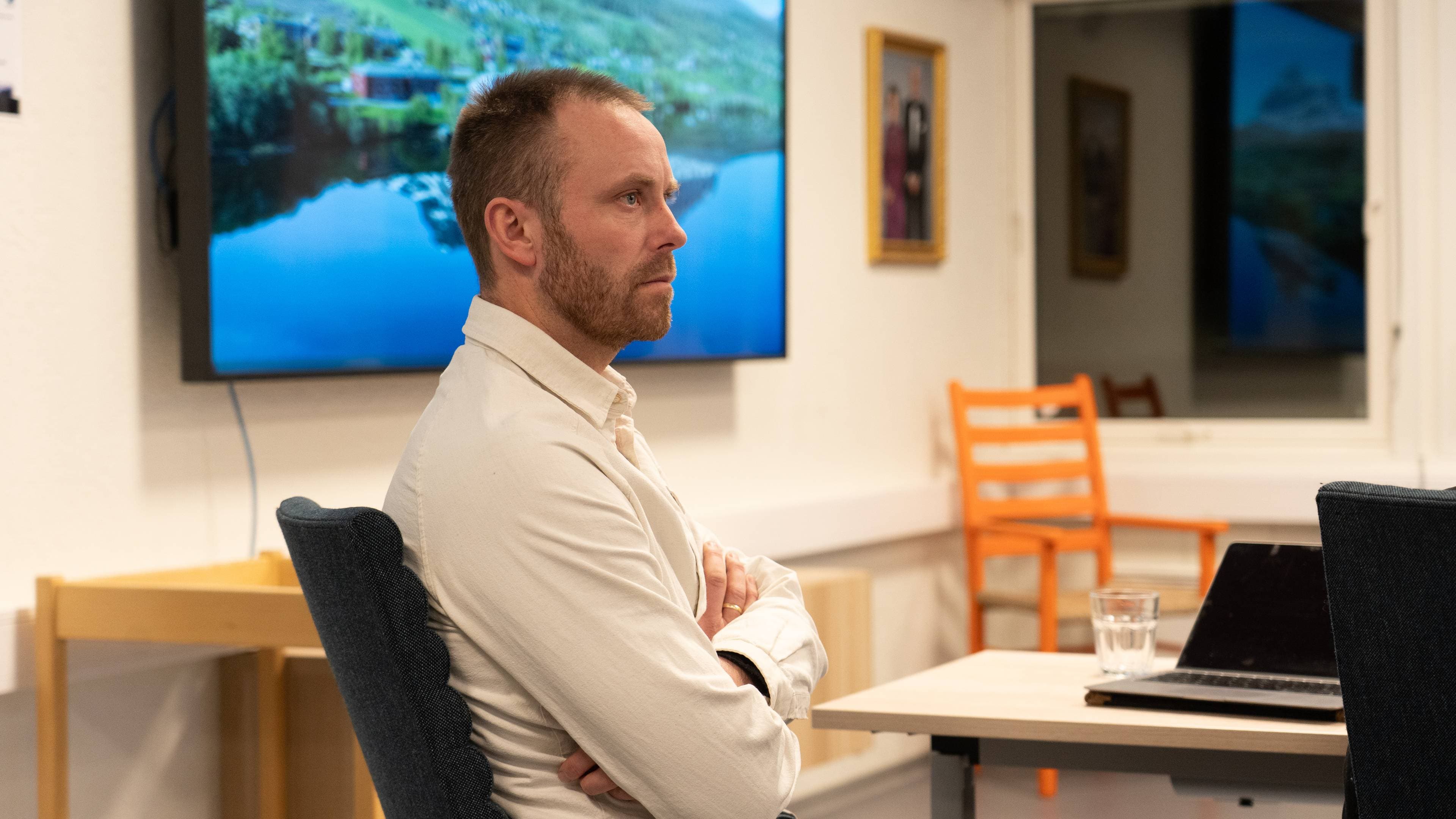 En person sitter med armene foldet i en stol, vendt mot en skjerm med landskapsbilde. I bakgrunnen er det et vindu og flere stoler, samt bilder på veggen. Et bord med en laptop og et glass vann er synlig foran personen. Rommet har en lys og enkel innredning.