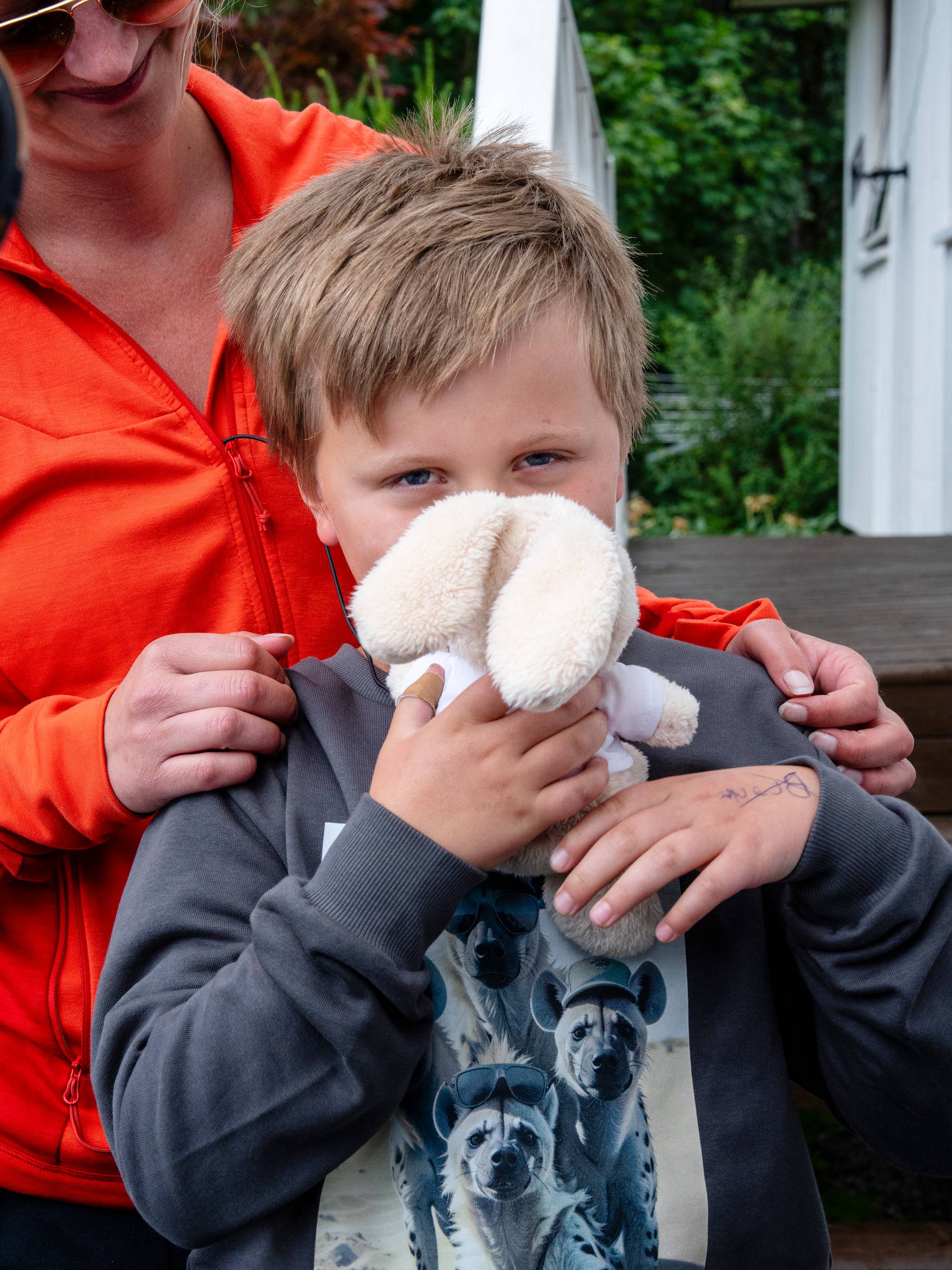 En kvinne i oransje jakke står bak en gutt som holder et kosedyr opp mot ansiktet. (Bildebeskrivelsen er laget av en KI-tjeneste)