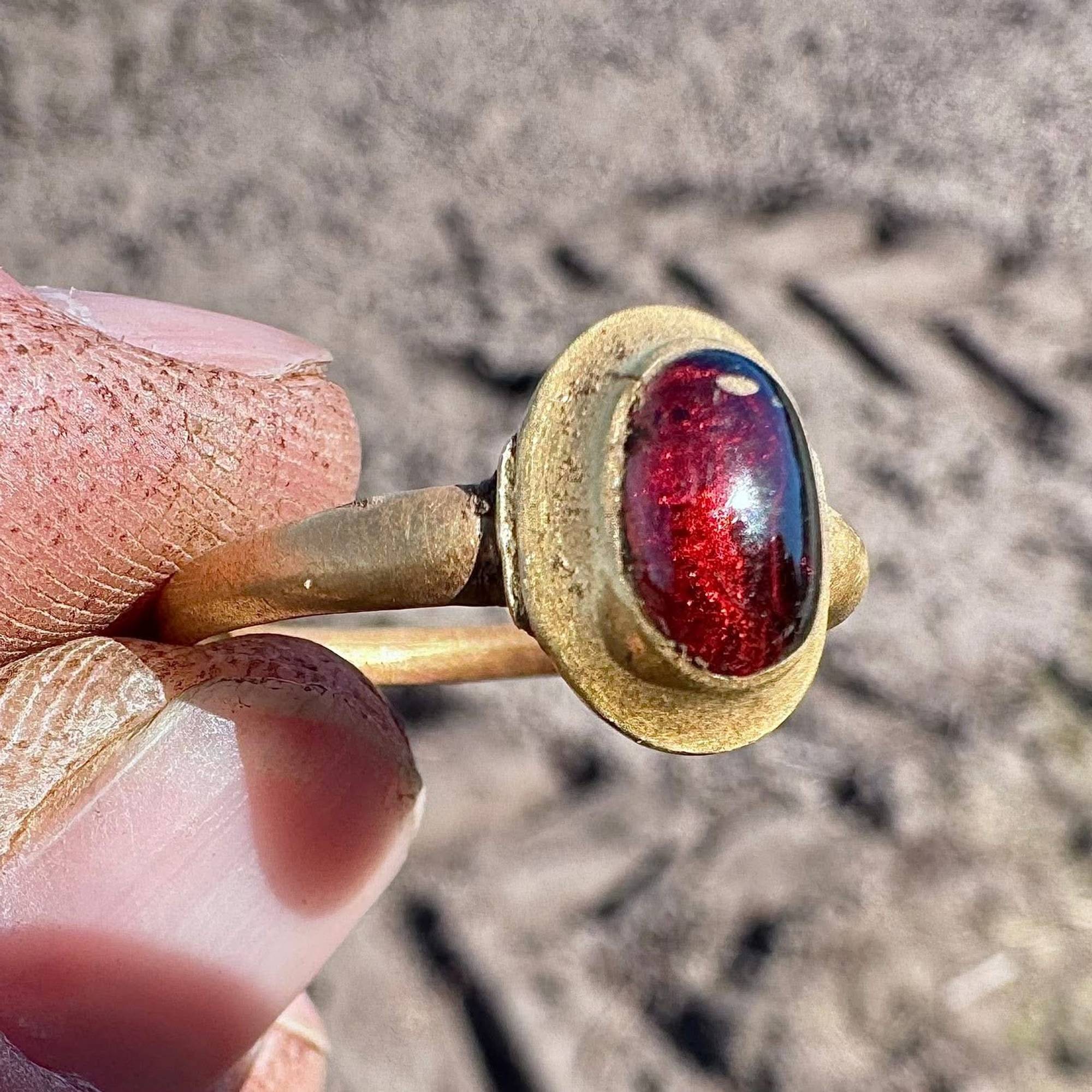 En person holder en ring med en rød stein. Ringen er av gull og har en oval steininnfatning. Bakgrunnen er uskarp og viser jord eller sand. Detaljer om oppdagelsen, som alder og funnsted, er ikke synlige i bildet. (Bildebeskrivelsen er laget av en KI-tjeneste)