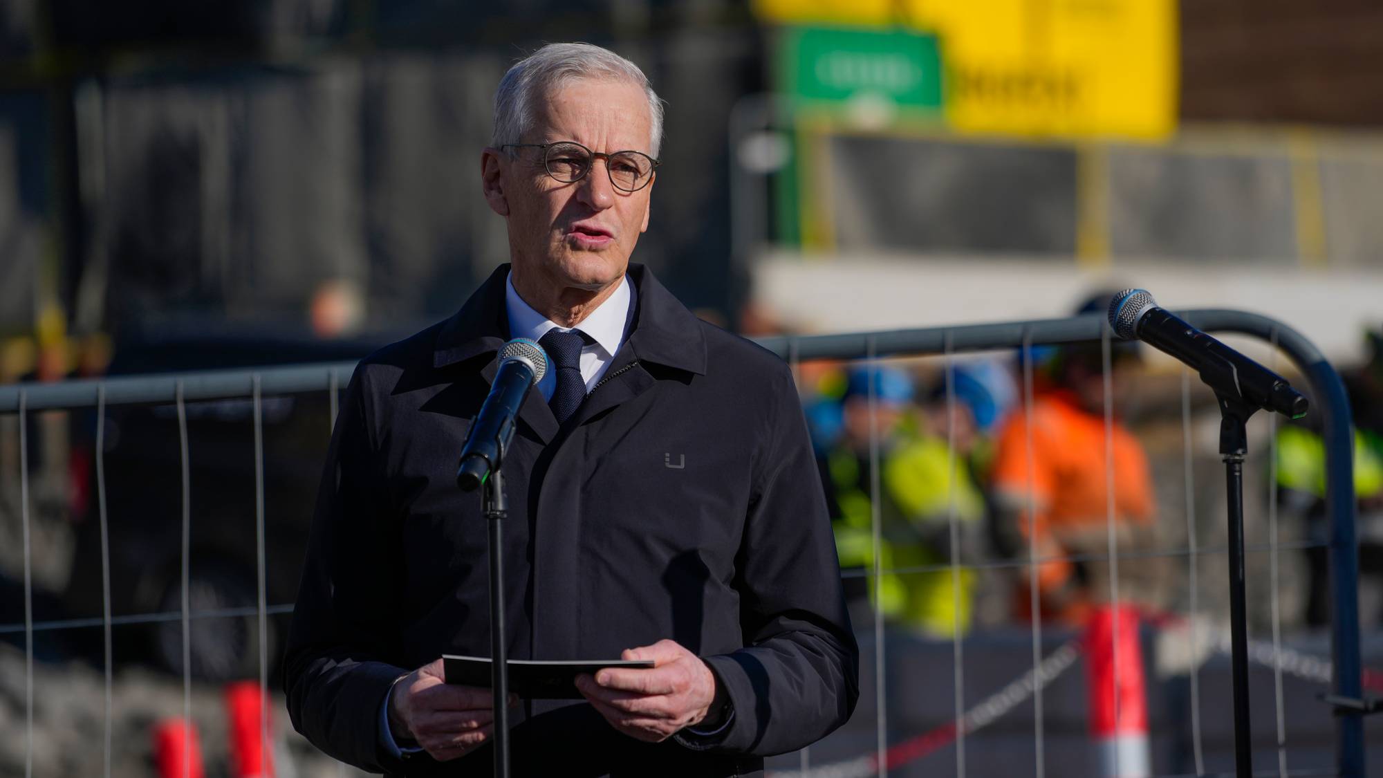 Jonas Gahr Støre snakker i en mikrofon. Han er kledd i mørk frakk og slips. Bak ham er et gjerde og uskarpe personer i gul og oransje vest. I bakgrunnen er det gule og grønne skilt. (Bildebeskrivelsen er laget av en KI-tjeneste)