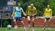 Fredrik Varfjell / NTB Fotballspillere på trening. Tre menn i treningstøy løper på en gressbane. En spiller foran er i lys blå drakt, og de to bak er i gul vest. I bakgrunnen er det en storskjerm og et gjerde. (Bildebeskrivelsen er laget av en KI-tjeneste)