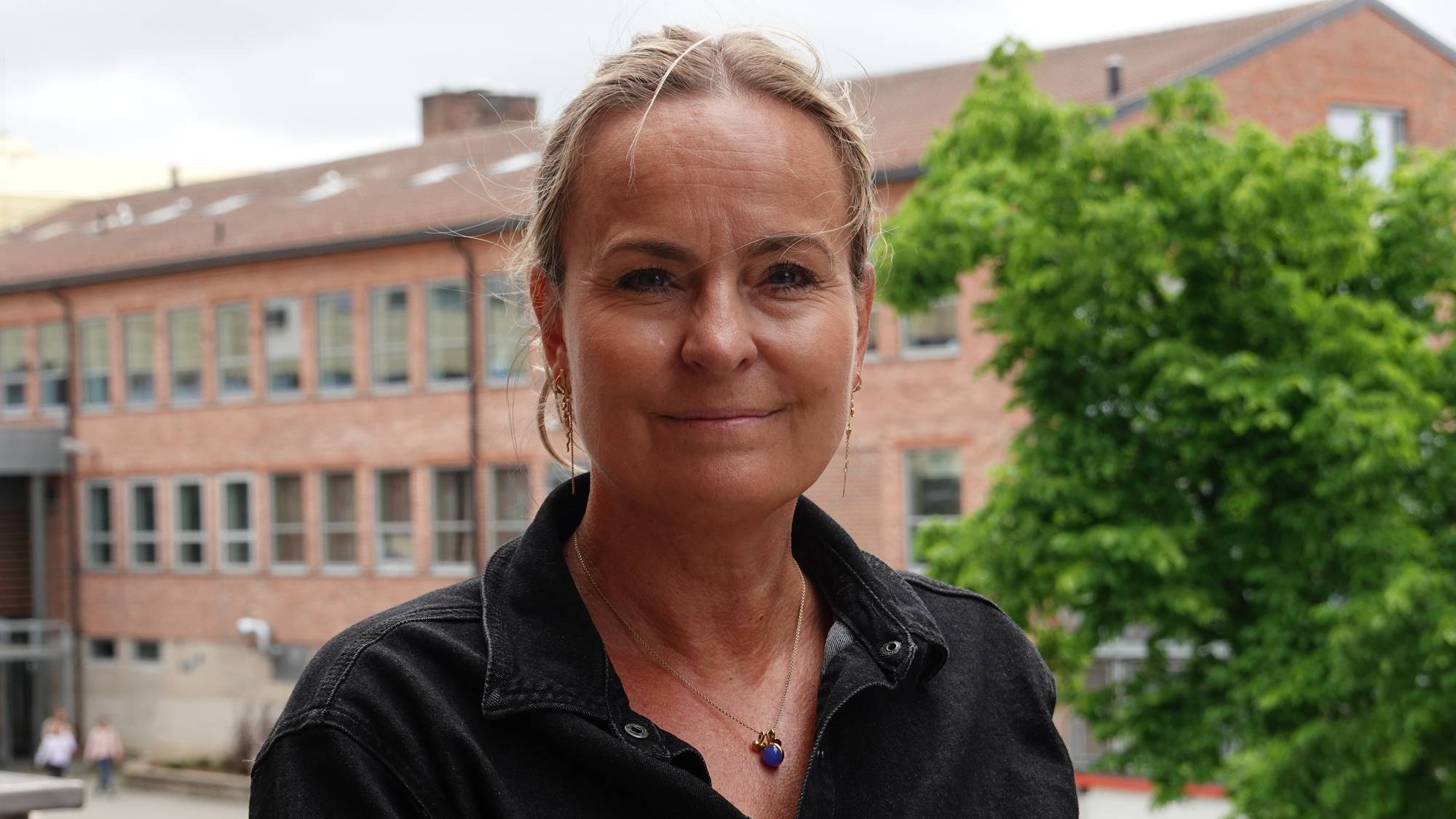 Portrett av en kvinne med blondt hår. Hun har på seg en mørk jakke og et smykke rundt halsen. I bakgrunnen ses en mursteinsbygning og et grønt tre. (Bildebeskrivelsen er laget av en KI-tjeneste)
