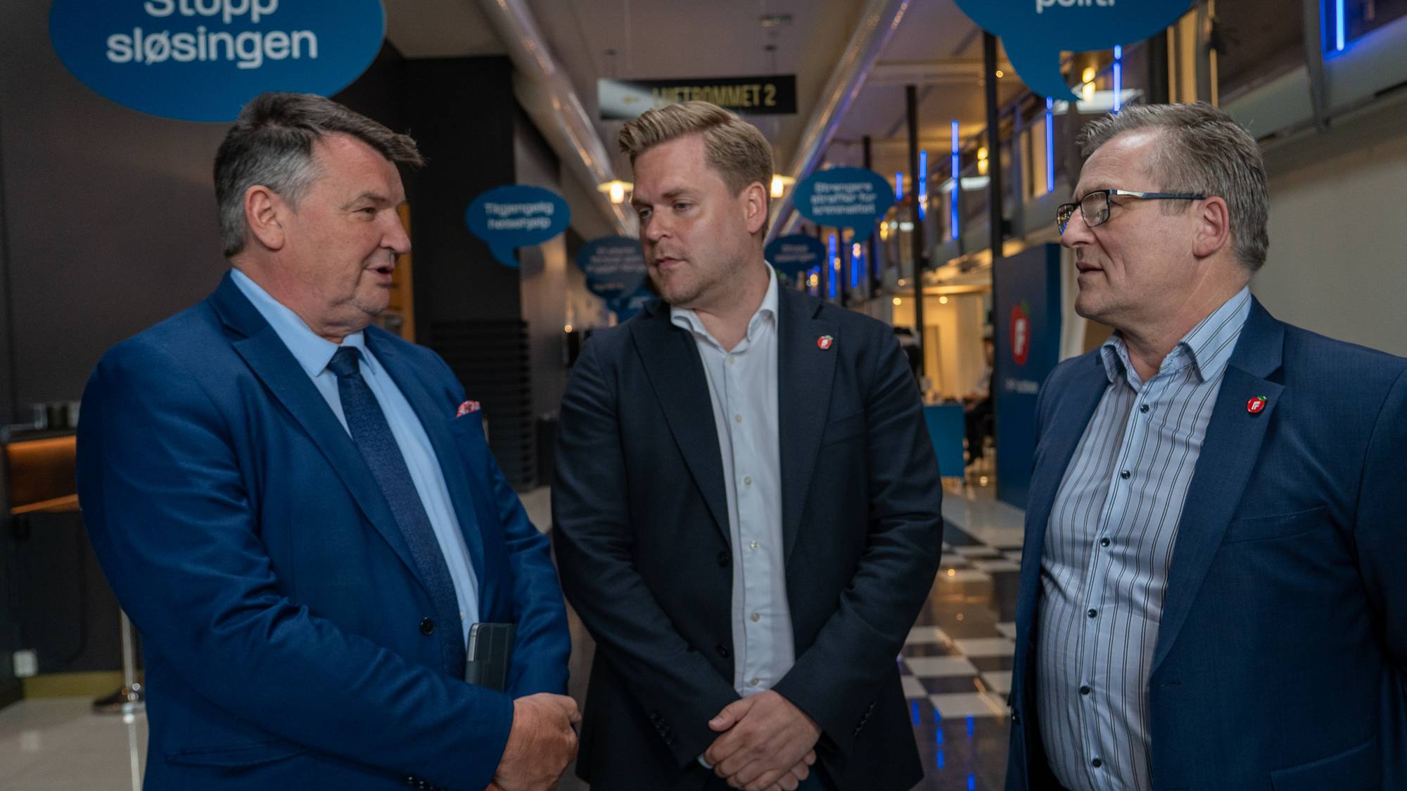 Tre menn i dress står og snakker sammen i en korridor. De har på seg slagord som "Stopp sløsingen" og "Mer synlig politi". Mennene ser ut til å være i en diskusjon. De har på seg dressjakker og skjorter. (Bildebeskrivelsen er laget av en KI-tjeneste)