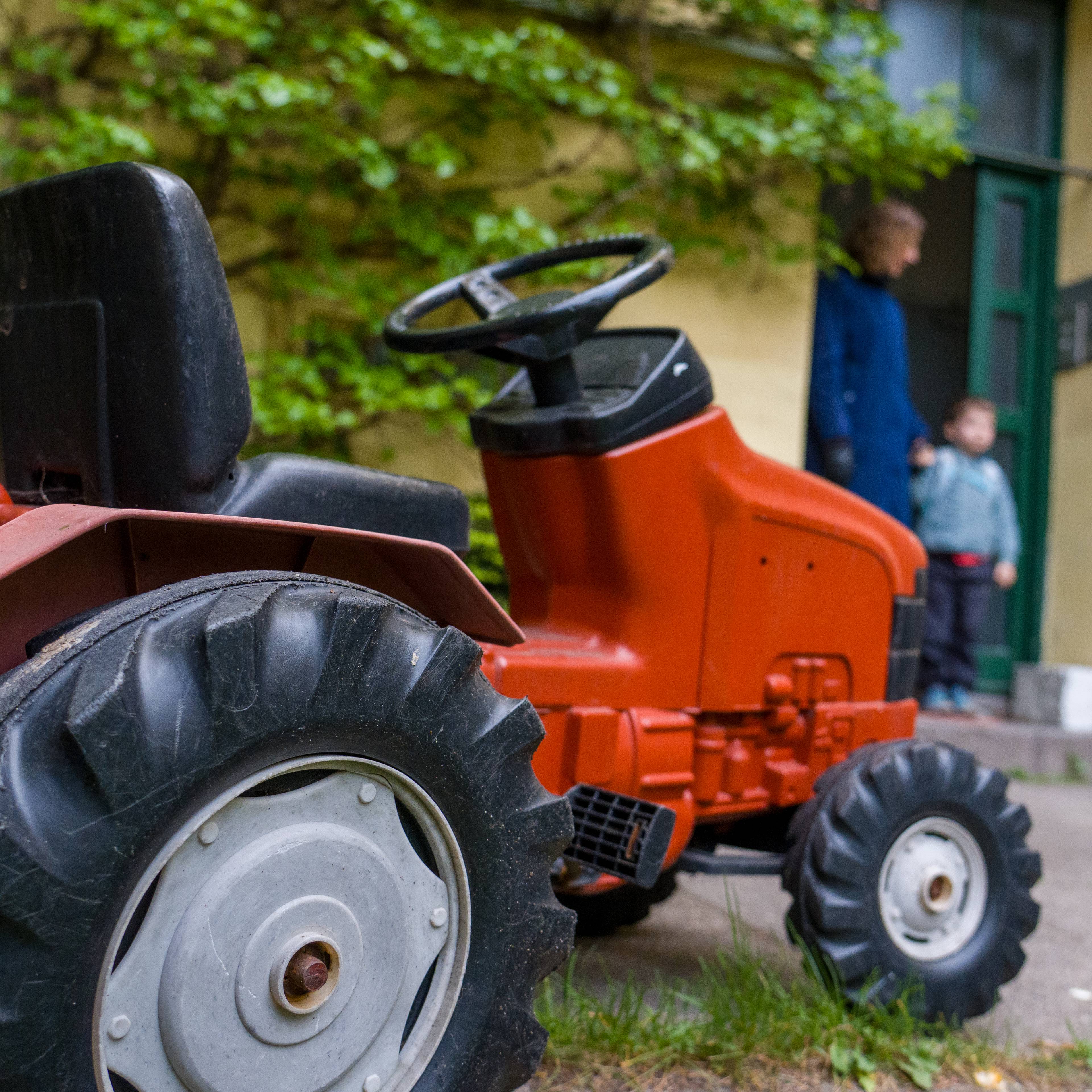 Bildet viser en rød leketraktor som står på en gressflekk. i bakgrunnen ser man en dame som holder en guttunge i hånden. De har begge på seg blå jakker.