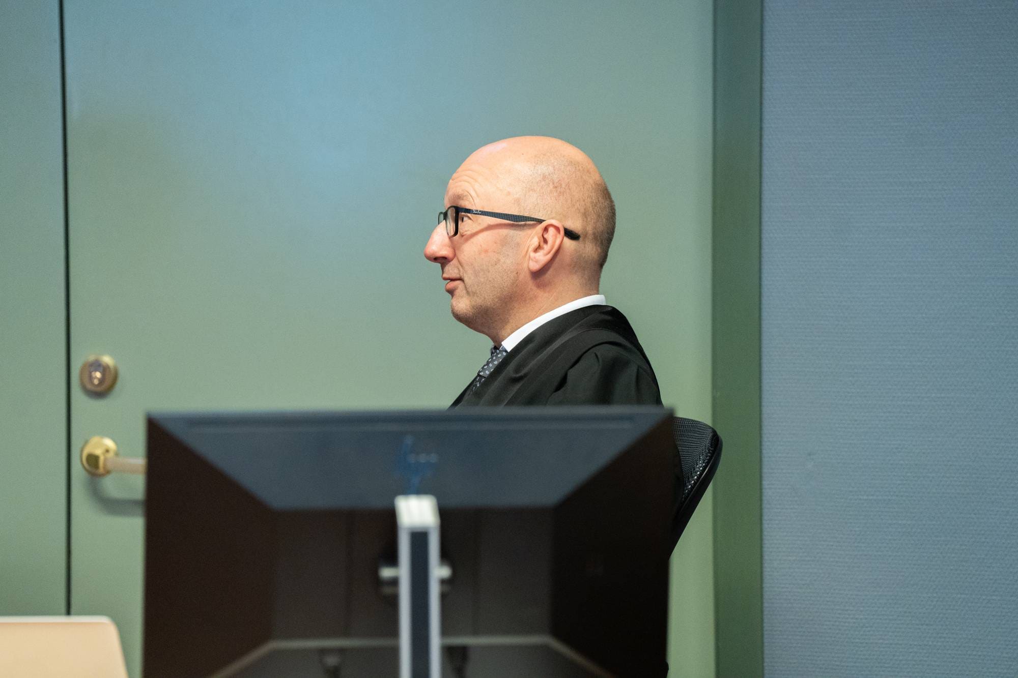 En mann i dress og kappe sitter foran en dataskjerm. Han har briller og er barbert på hodet. Mannen sitter i et rom med lysegrønne og blå vegger.