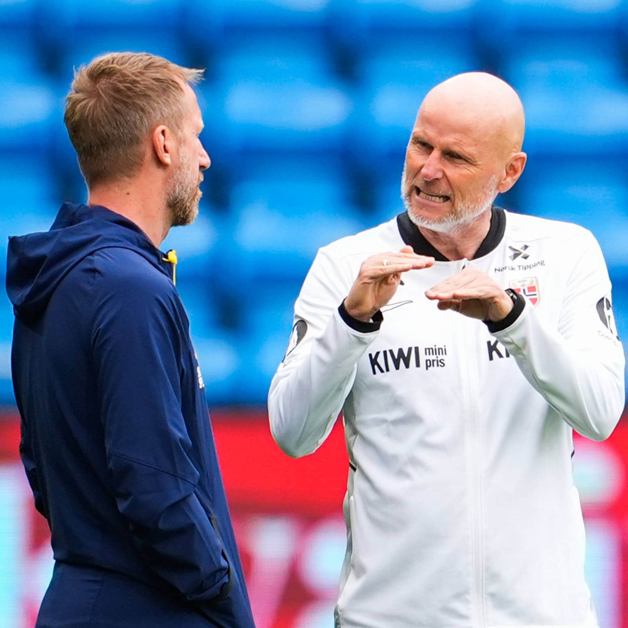 To trenere diskuterer på en fotballbane. Den ene med skallet hode, iført en hvit jakke, forklarer tydelig med hendene. Den andre, med kort, blondt hår, lytter med siden til. I bakgrunnen sees blå tribunefelt. (Bildebeskrivelsen er laget av en KI-tjeneste)