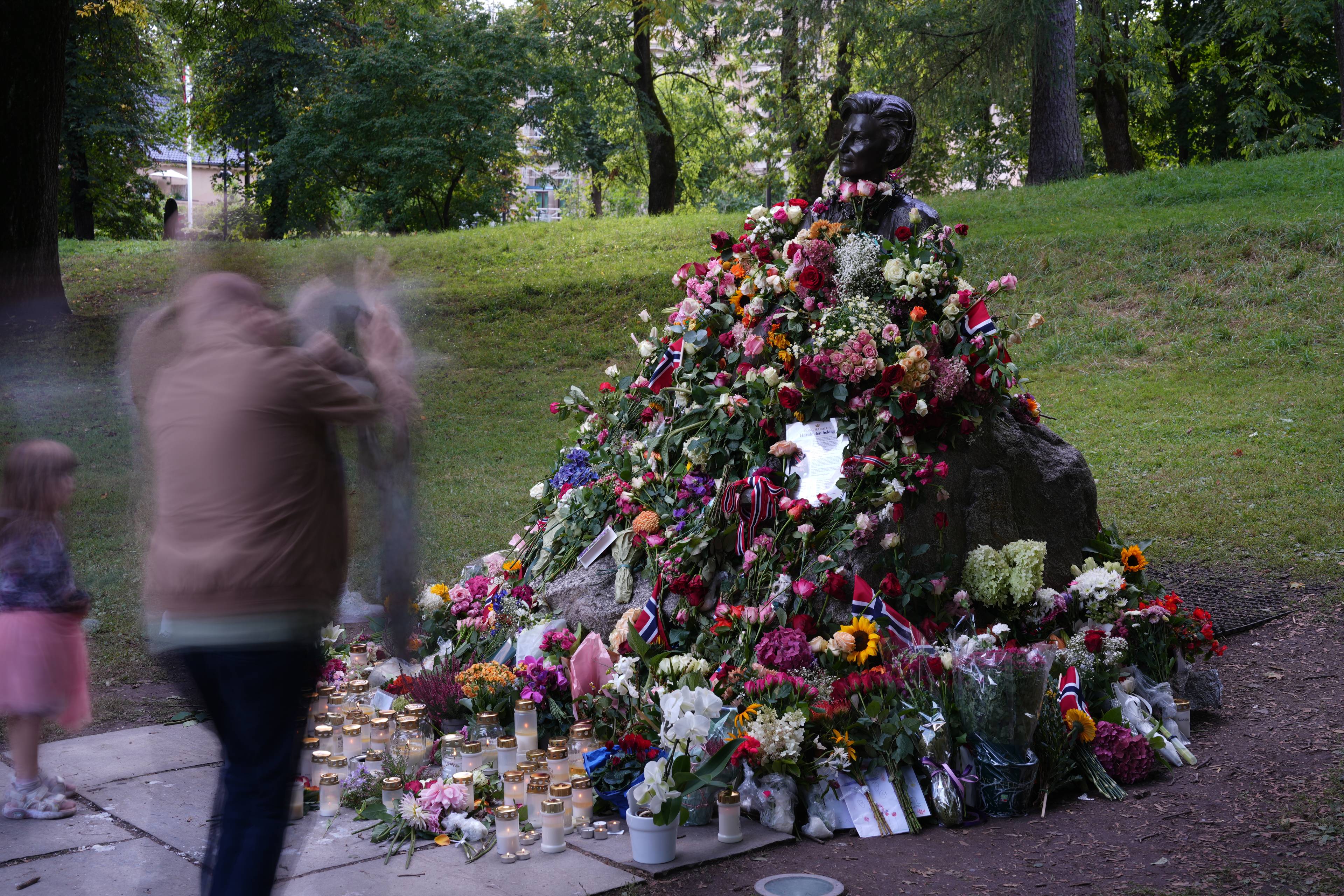 En byste dekket av blomster og norske flagg i en park, med tente lys og besøkende foran. (Bildebeskrivelsen er laget av en KI-tjeneste)