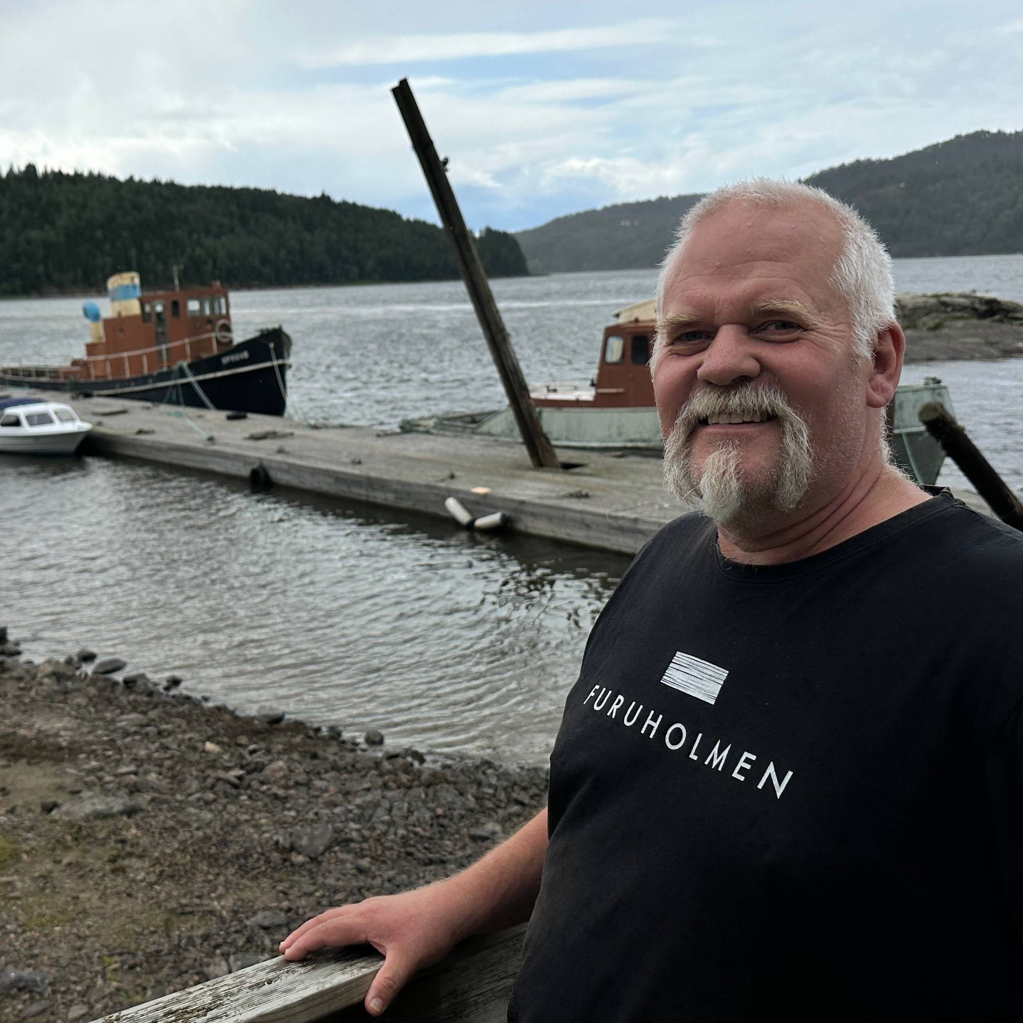 Mann med grått skjegg ved en brygge med båter i en innsjø. (Bildebeskrivelsen er laget av en KI-tjeneste)