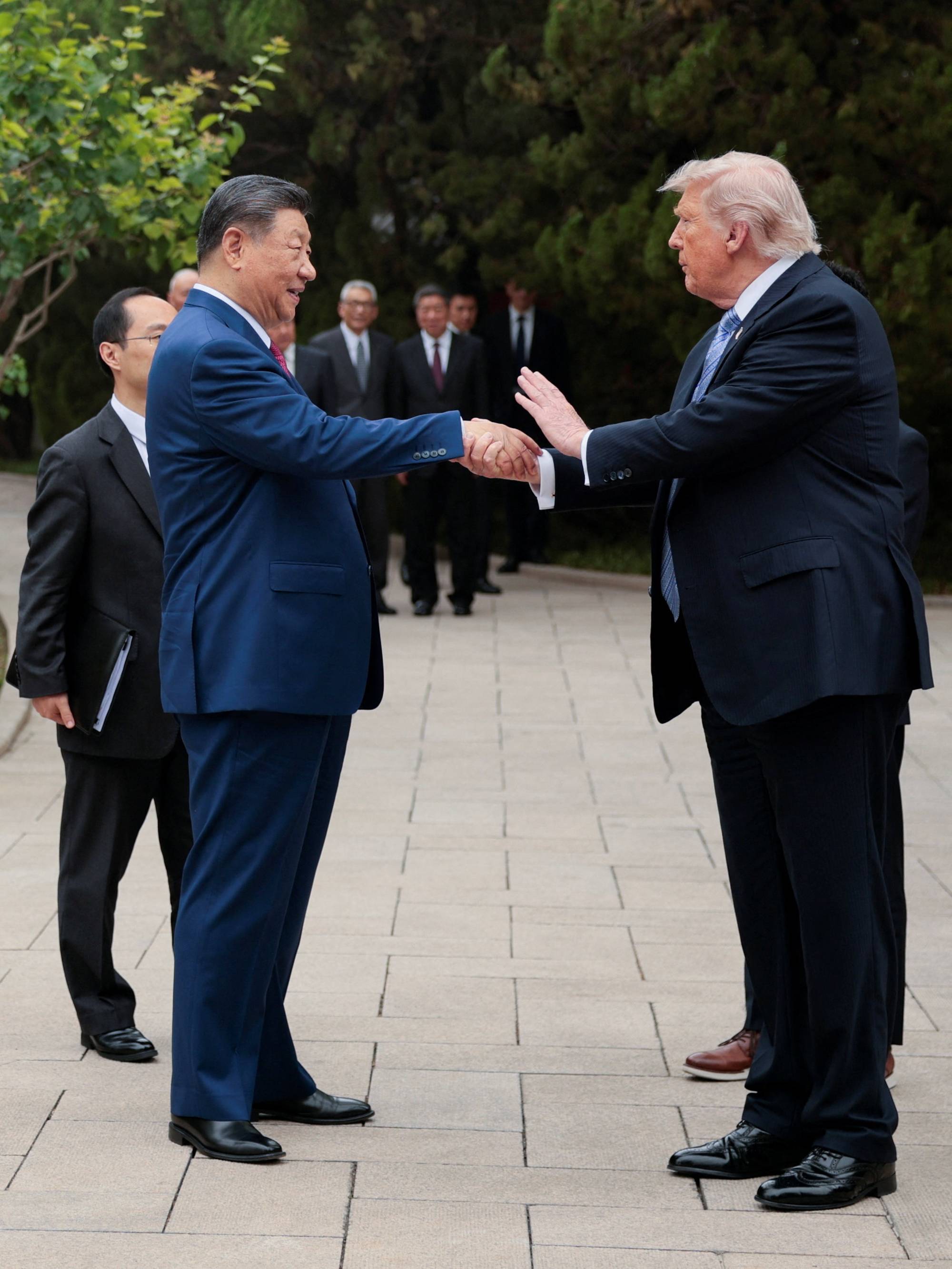 Kinas president Xi Jinping hilser på USAs president Donald Trump. De to lederne står utendørs og håndhilser. Flere menn i dress står bak dem. (Bildebeskrivelsen er laget av en KI-tjeneste)