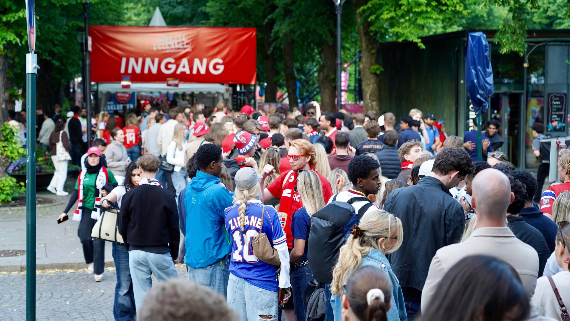 En stor folkemengde samles ved en rød INNGANG-banner. Mange bærer supporterutstyr, inkludert landslagsdrakter og hodeplagg. Stemningen virker forventningsfull, og det er sannsynligvis et arrangement eller en kamp som nærmer seg. Omgivelsene preges av grønne trær og en brosteinsbelagt gate. (Bildebeskrivelsen er laget av en KI-tjeneste)