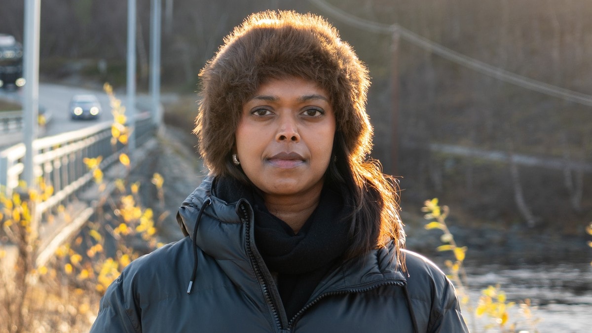 Kvinne med pelskantet lue og dunjakke står ved en veg med ei bru i bakgrunnen. (Bildebeskrivelsen er laget av en KI-tjeneste) FOTO: Kristina Kalinina/NRK
