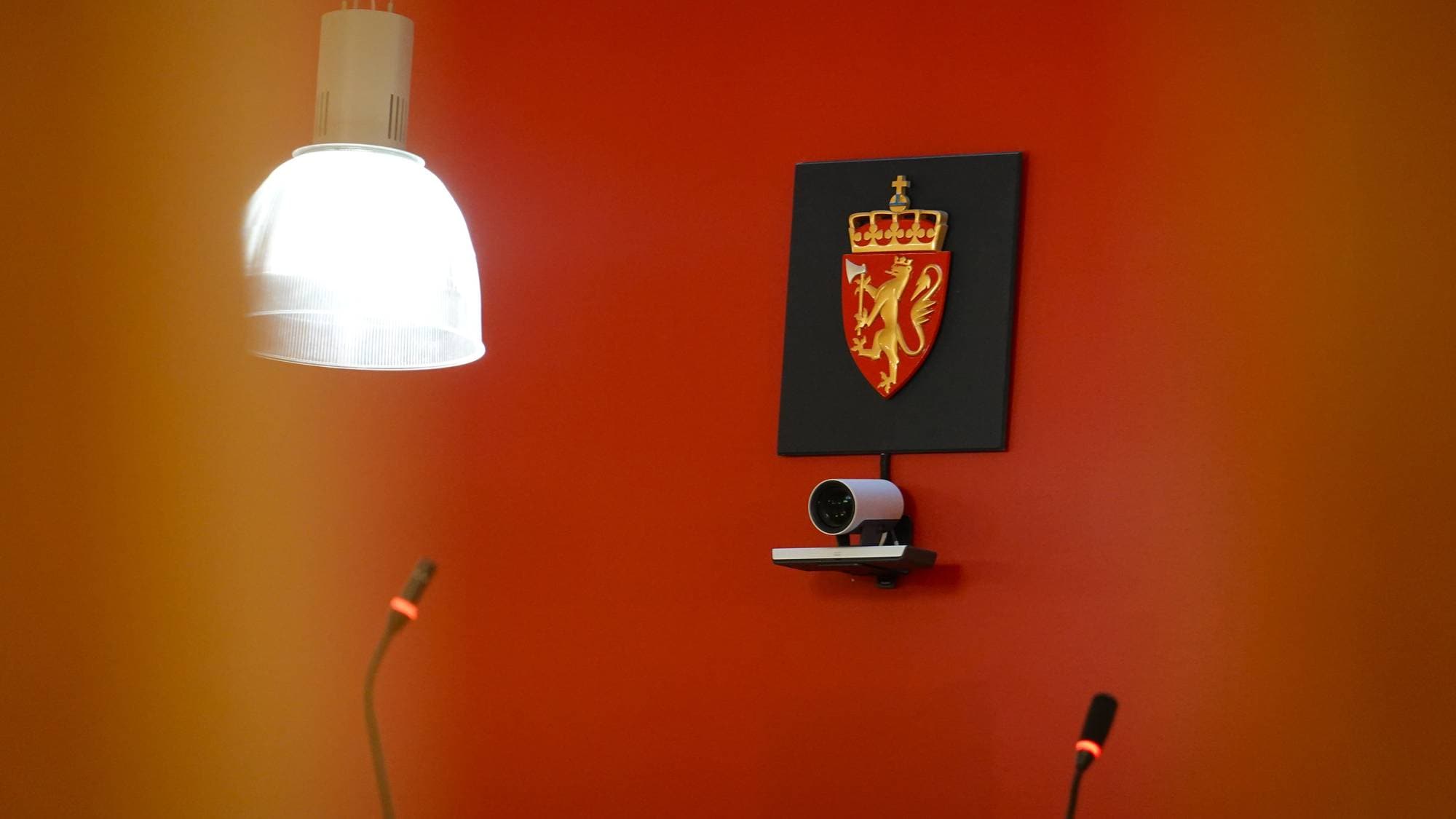 Det norske riksvåpenet henger på en rød vegg over et kamera, flankert av en taklampe og mikrofoner. (Bildebeskrivelsen er laget av en KI-tjeneste)