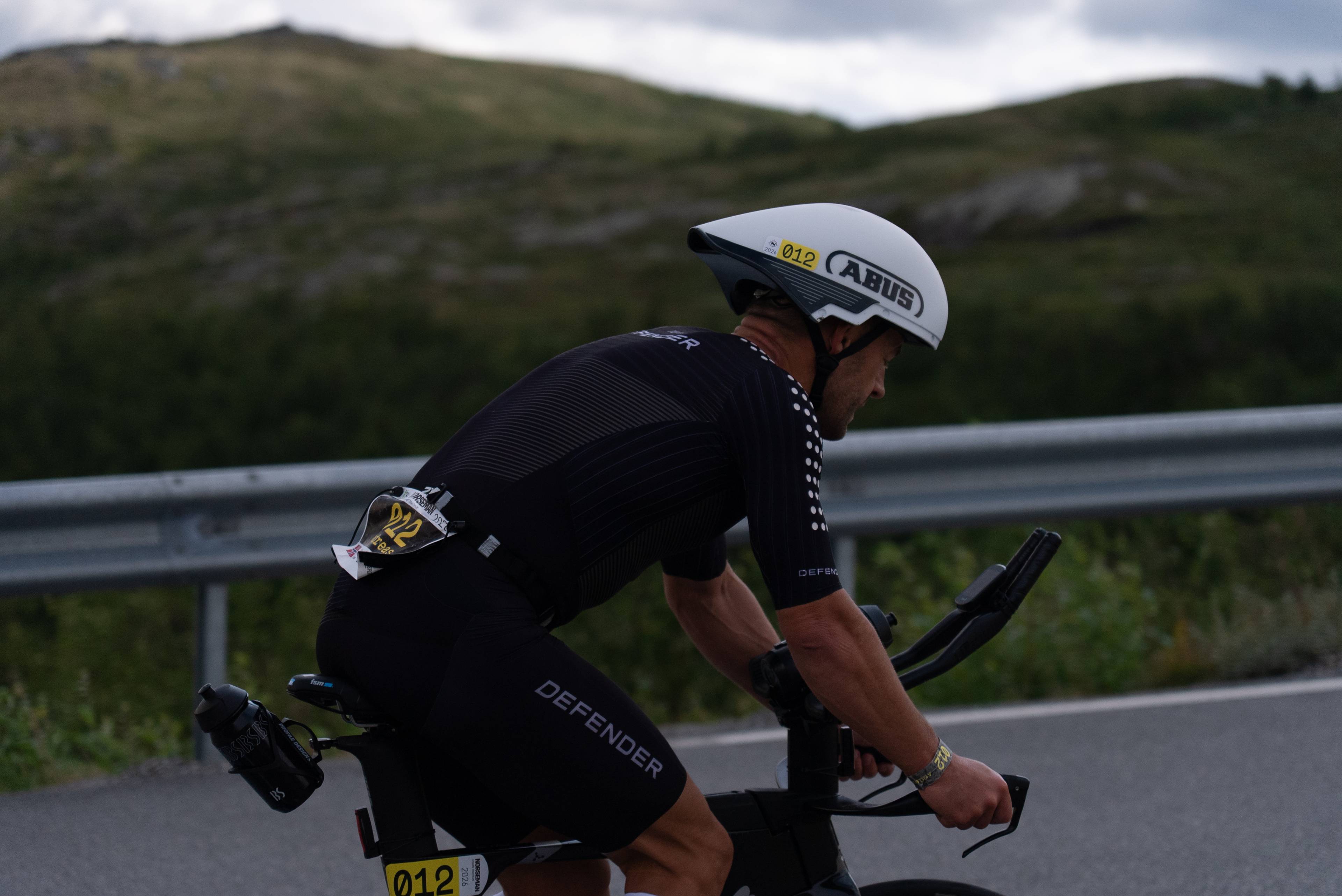 Syklist i triatlonkonkurranse med hjelm og startnummer sykler langs en vei i fjellterreng. (Bildebeskrivelsen er laget av en KI-tjeneste)