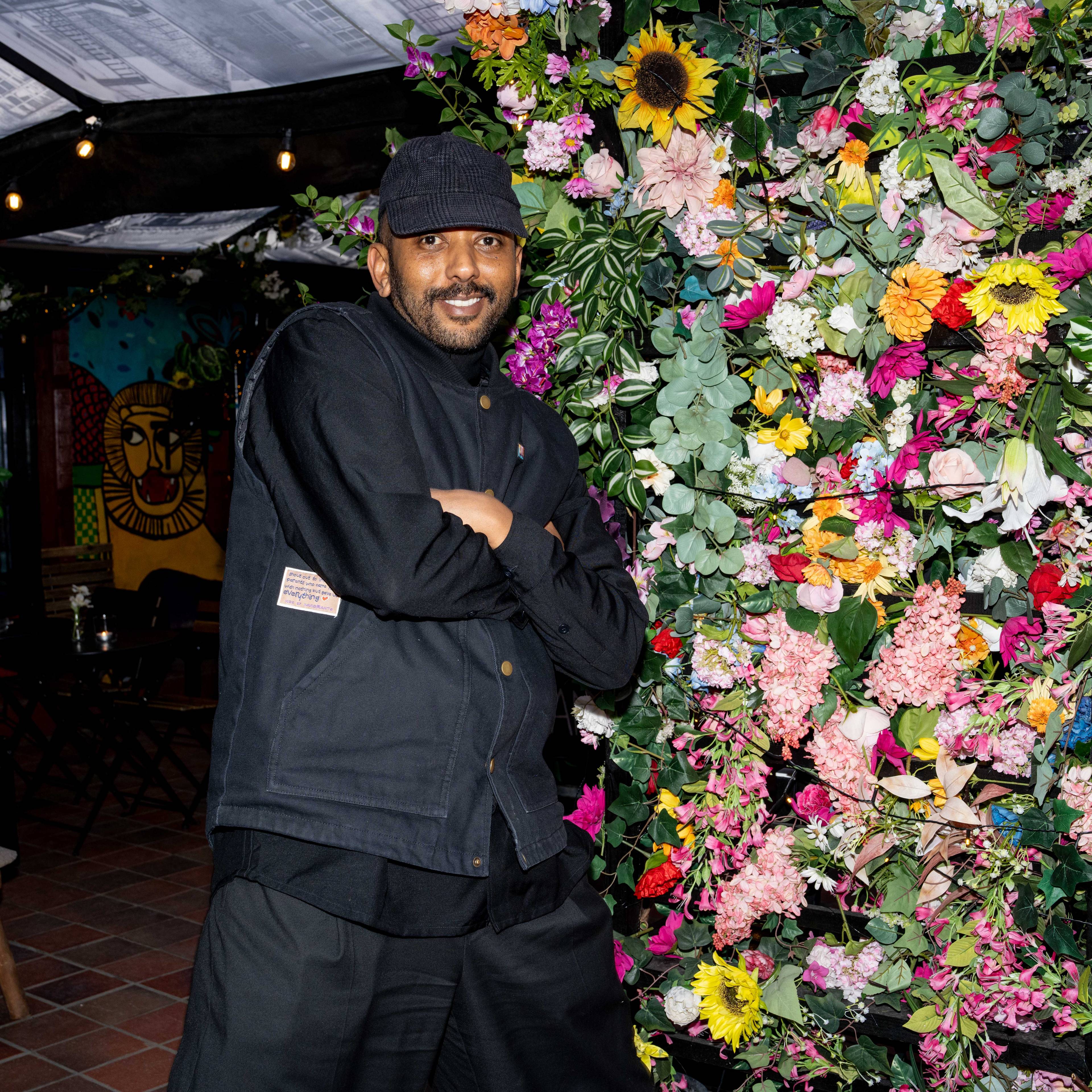 Misgina poserer med armene i kryss og smiler. I bakgrunnen ser vi uteservering til en bar, og til høyre for Misgina er en vegg med blomster i alle slags farger.